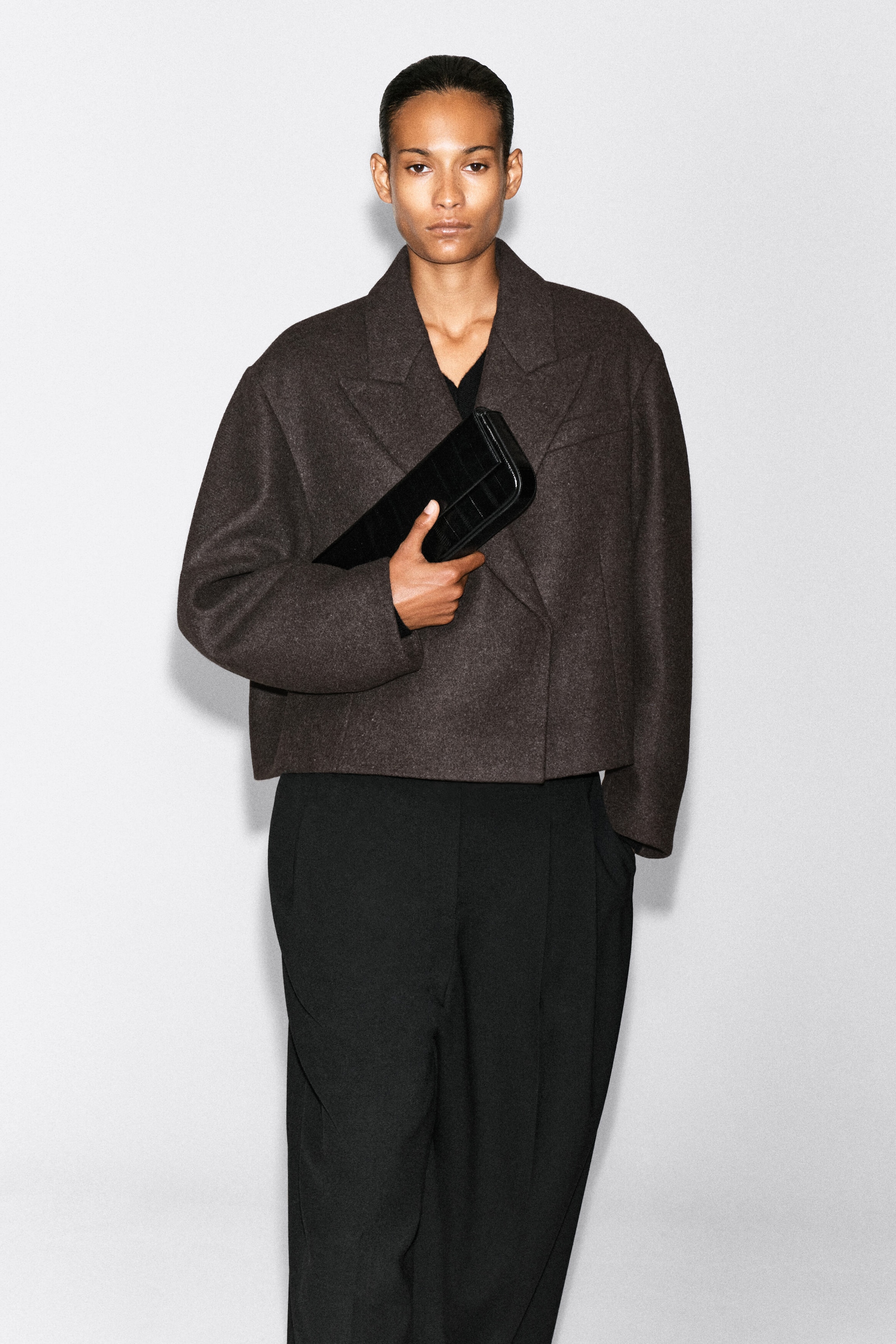 An oversized, cropped boiled-wool blend blazer in dark brown is worn by the model, featuring notch lapels, dropped shoulders, and a textured finish. The blazer is paired with black wide-leg trousers, and the model is holding a black textured clutch bag across the front. An oversized, cropped boiled-wool blend blazer in dark brown is worn by the model, featuring notch lapels, dropped shoulders, and a textured finish. The blazer is paired with black wide-leg trousers, and the model is holding a black textured clutch bag across the front.