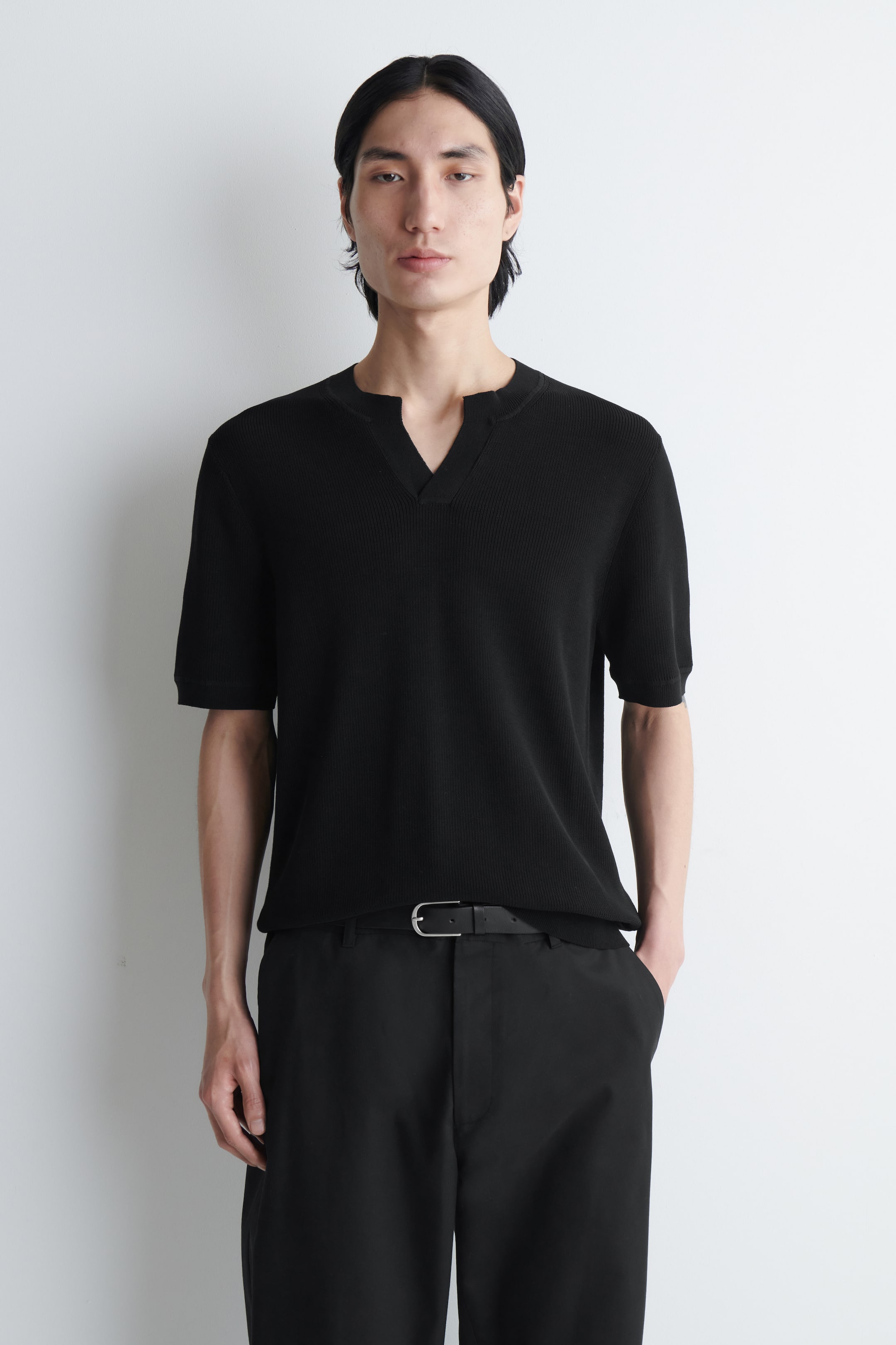 Vezi imaginea mai mare: Modelul poartă un tricou polo negru cu mânecă scurtă, tricotat, cu o textură în relief și un decolteu în V deschis. Modelul stă în picioare, purtând tricoul alături de pantaloni negri și o curea neagră.