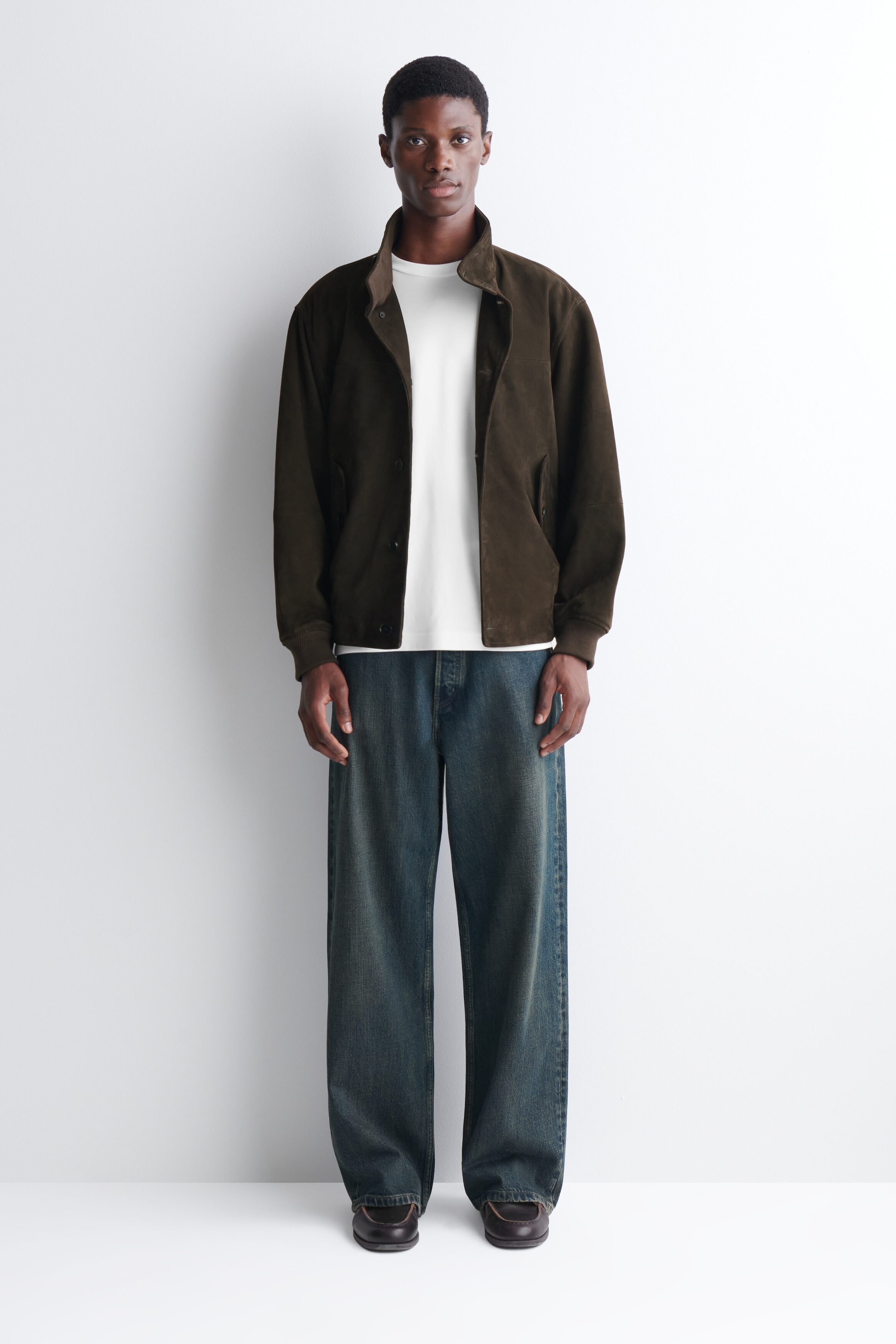A dark brown suede jacket is being worn open by the model, featuring a stand collar, front button fastenings, and ribbed cuffs. The jacket is layered over a plain white crew-neck T-shirt and paired with loose-fitting, dark-wash blue jeans. A dark brown suede jacket is being worn open by the model, featuring a stand collar, front button fastenings, and ribbed cuffs. The jacket is layered over a plain white crew-neck T-shirt and paired with loose-fitting, dark-wash blue jeans.