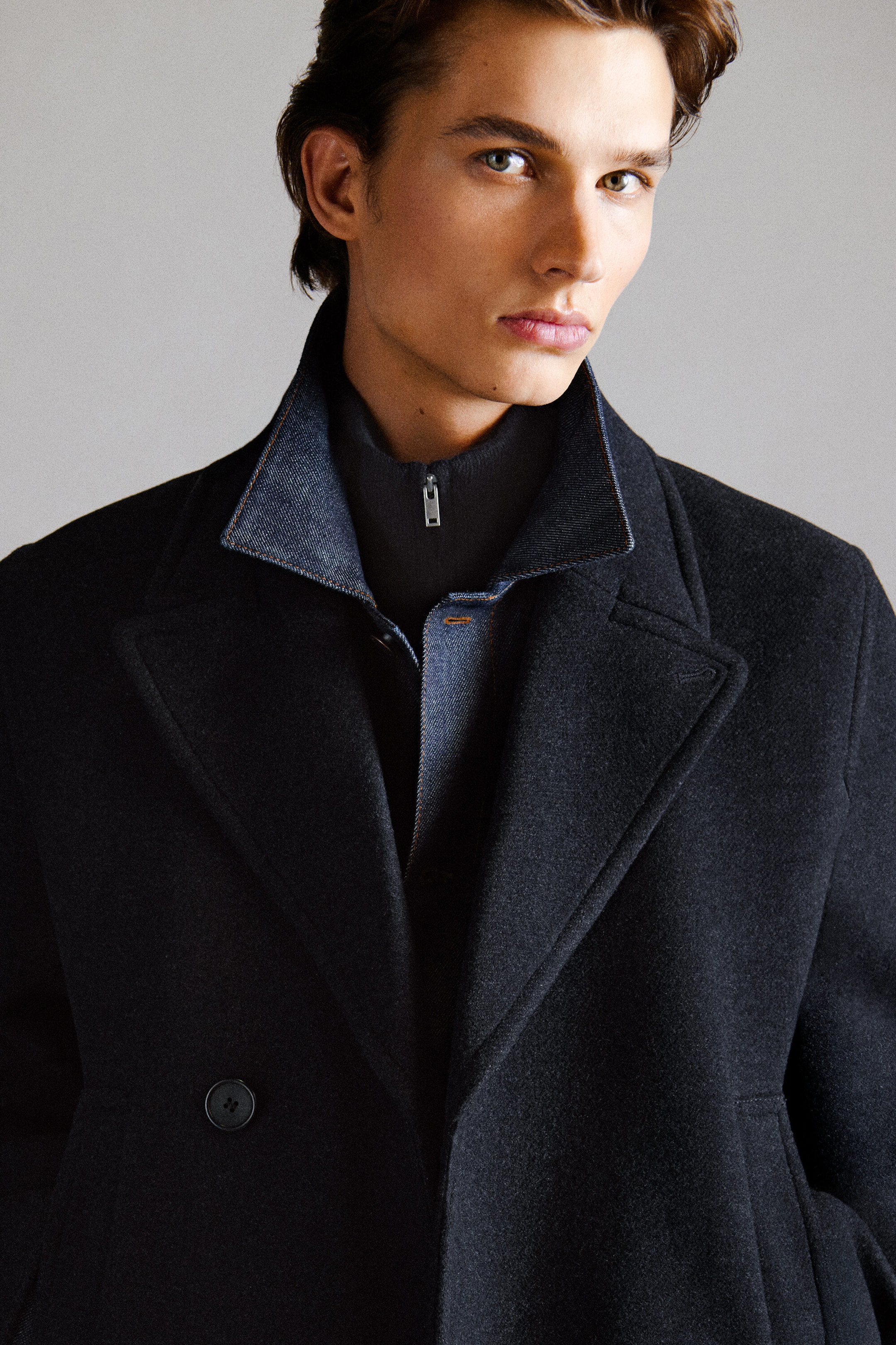 A dark grey double-breasted wool pea coat is shown in a front-facing close-up, highlighting its wide notch lapels and structured fit. The coat is layered over a blue denim shirt with an upturned collar and a black zip-up high-neck sweater.