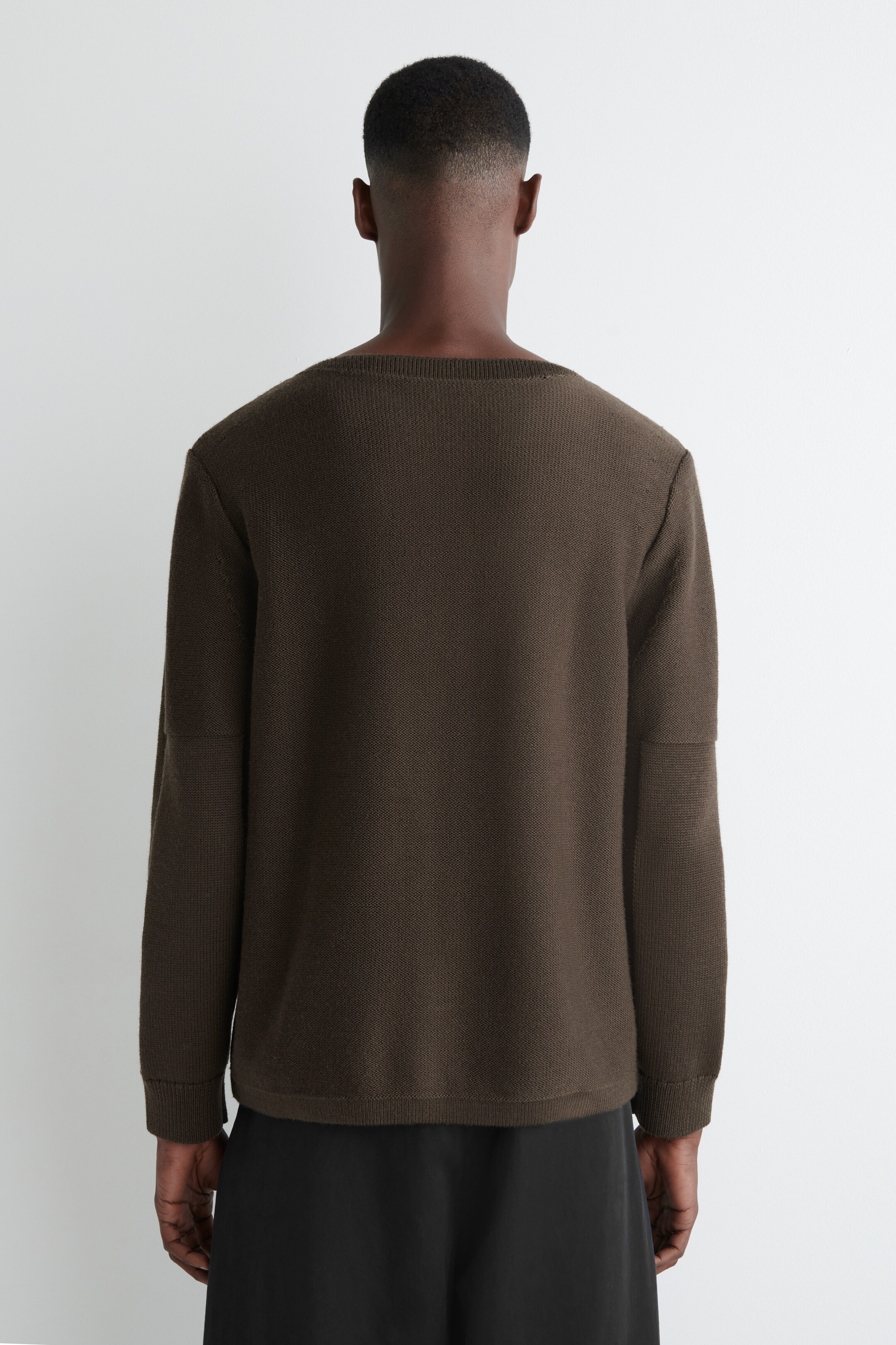 Agrandir l'image: Un pull en tricot marron foncé est vu de dos, avec une coupe décontractée, un large col peu profond et des manches longues avec un détail de couture horizontale sur le haut du bras. L'ourlet droit du pull repose au-dessus d'un pantalon noir.