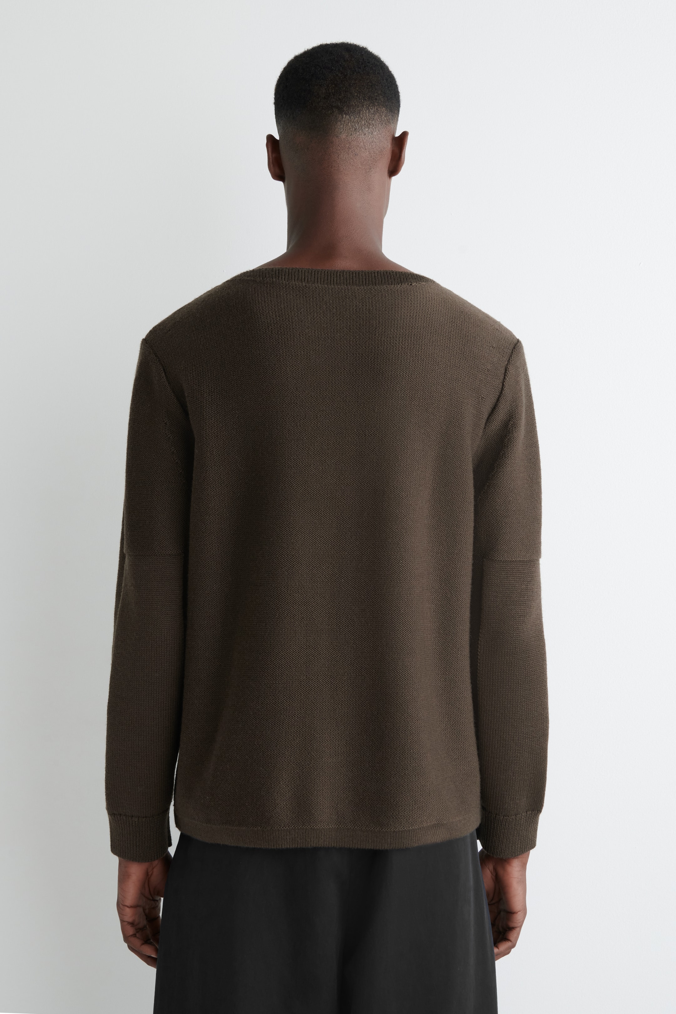 Grösseres Bild ansehen: Ein dunkelbrauner Strickpullover wird von hinten betrachtet, mit einer entspannten Passform, einem breiten, flachen Ausschnitt und langen Ärmeln mit einem horizontalen Nahtdetail am Oberarm. Der gerade Saum des Pullovers ruht über schwarzen Hosen.