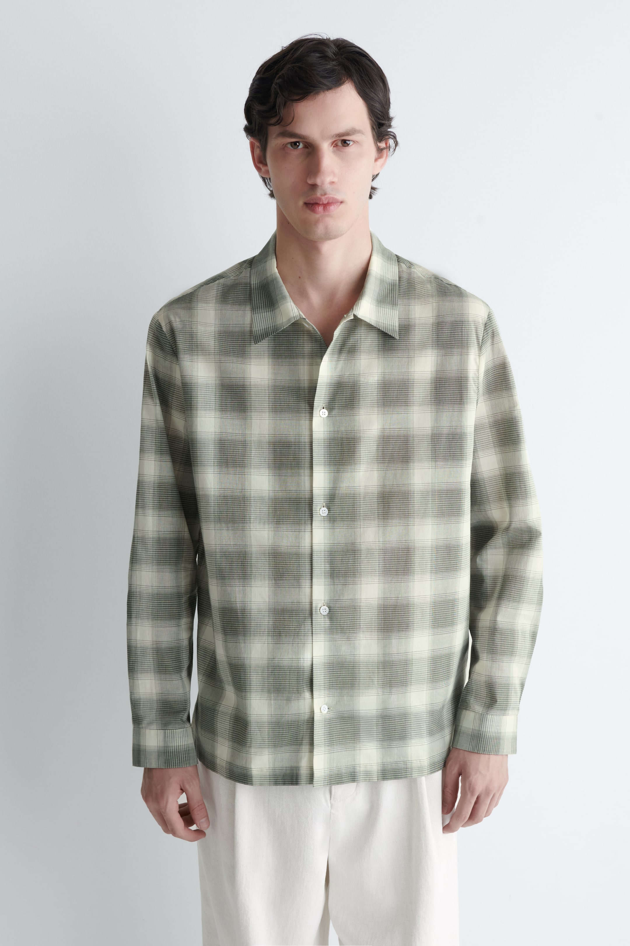 Agrandir l'image: Une chemise à manches longues est exposée, face avant, arborant un motif à carreaux dans des tons vert olive et crème, avec des textures de rayures horizontales subtiles à l'intérieur des carreaux plus foncés. Elle dispose d'un col évasé, d'une patte de boutonnage sur le devant avec des boutons blancs, et de poignets simples.