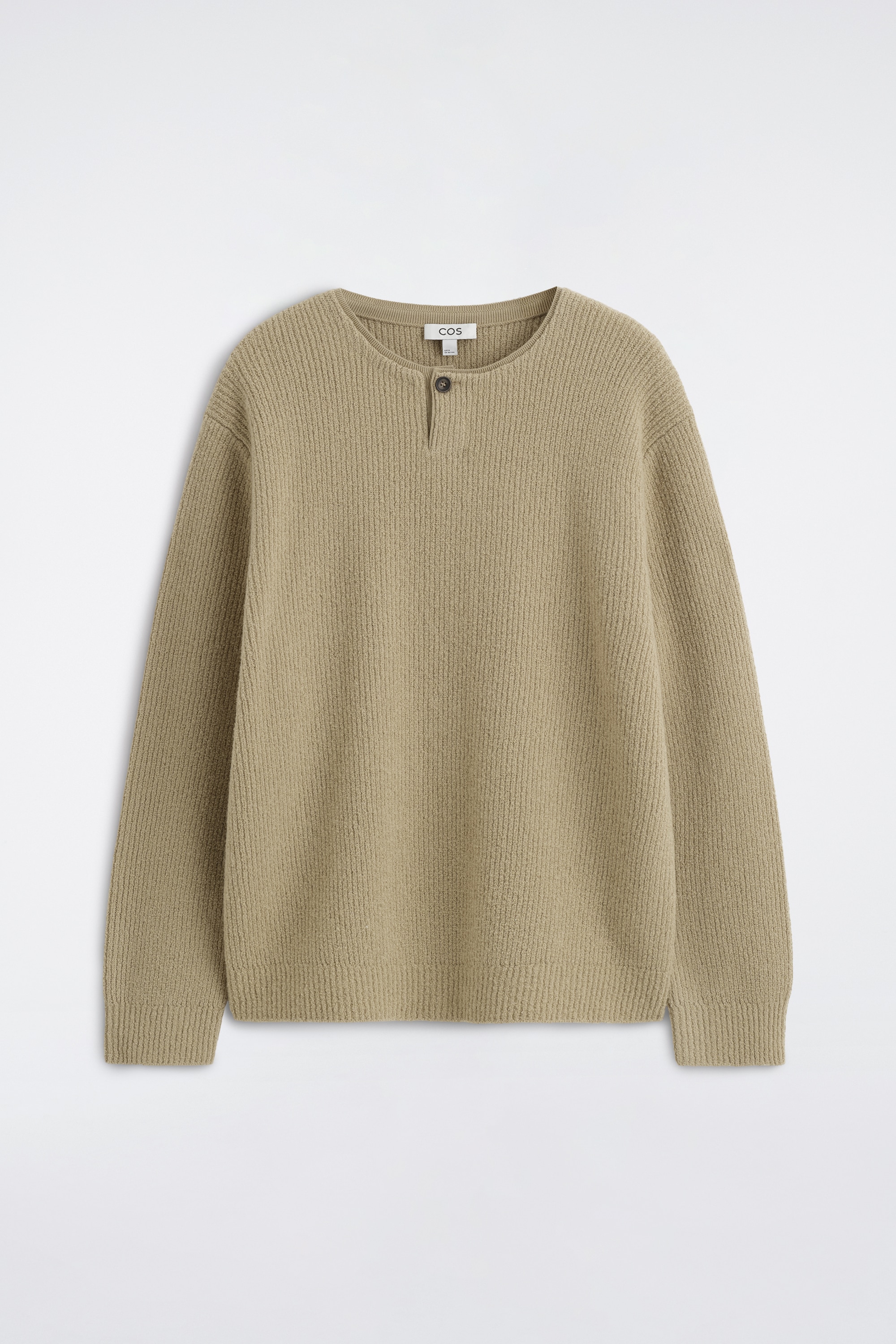 Een beige henleytrui in ribgebreid materiaal wordt gedragen door het model, met een sluiting met Ã©Ã©n knoop aan de kraag en een losse pasvorm. De trui met lange mouwen is gecombineerd met donkerblauwe pantalon.