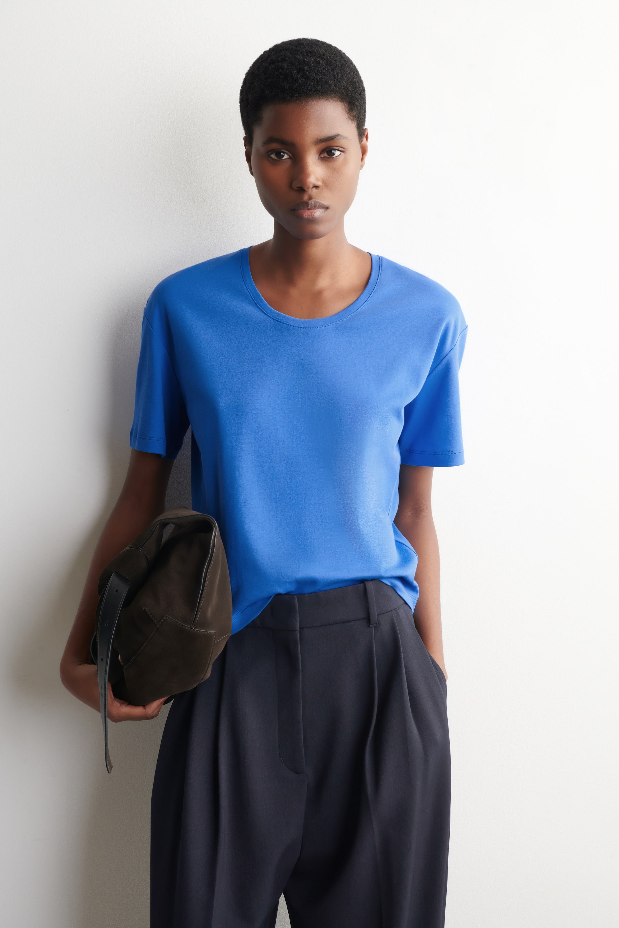 Größeres Bild ansehen: Ein leuchtend blaues T-Shirt mit einem tiefen Ausschnitt, überschnittenen Schultern und einer lockeren Passform wird vom Model getragen. Das Model kombiniert das T-Shirt mit dunklen Hosen und hält eine dunkle Tasche.