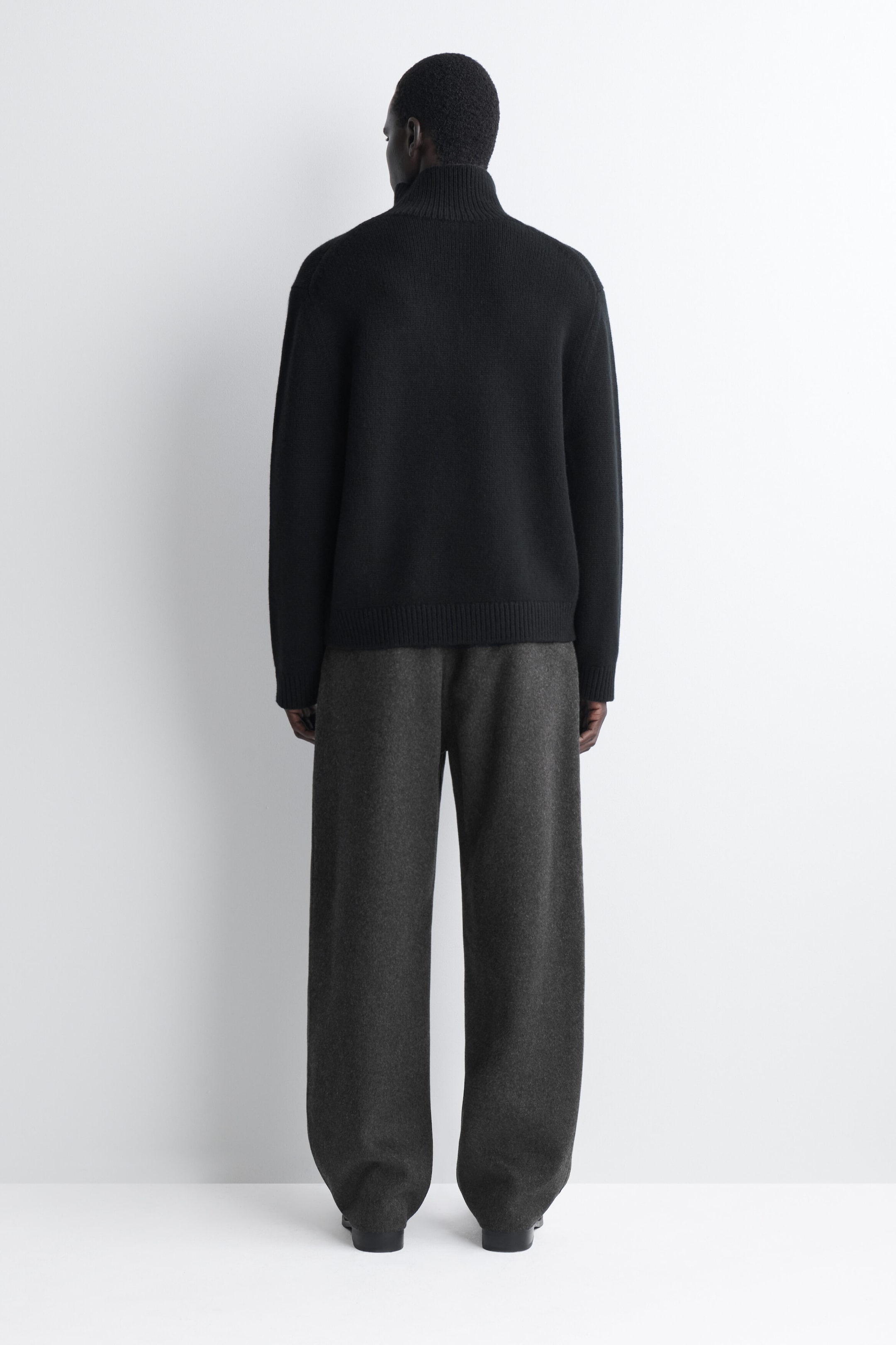 Ein schwarzer Strickpullover ist von hinten zu sehen. Er hat einen hohen, gerippten Stehkragen, gerippte Bündchen und einen gerippten Saum und wird mit dunkelgrauen, weit geschnittenen Hosen kombiniert. Ein schwarzer Strickpullover ist von hinten zu sehen. Er hat einen hohen, gerippten Stehkragen, gerippte Bündchen und einen gerippten Saum und wird mit dunkelgrauen, weit geschnittenen Hosen kombiniert.