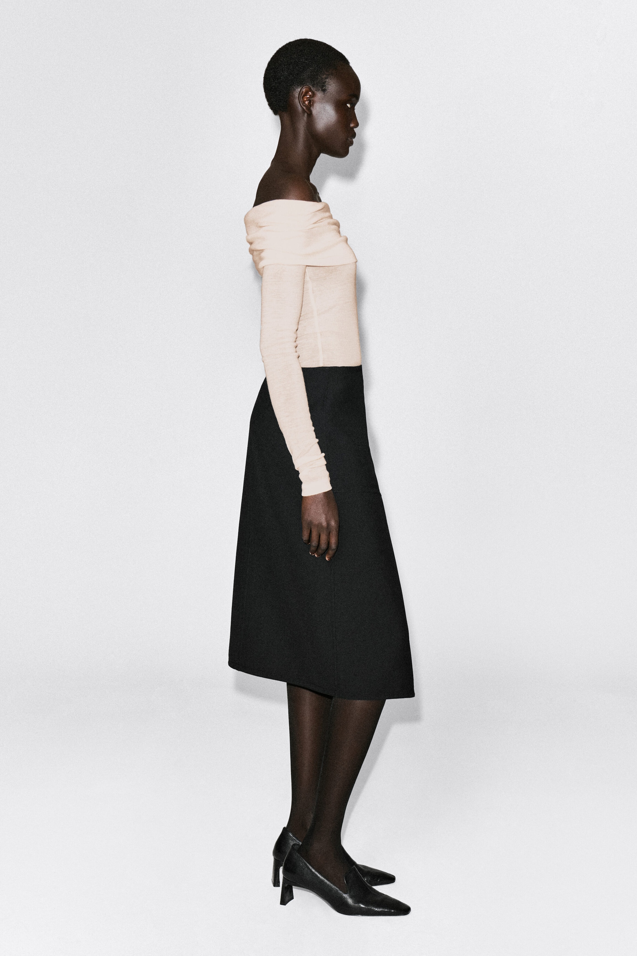 An off-the-shoulder merino wool top in a pale cream shade is shown in a right-facing side profile, highlighting its folded neckline and slim-fitting long sleeves. The lightweight, semi-sheer top is tucked into a high-waisted black skirt, emphasizing its close fit along the torso. An off-the-shoulder merino wool top in a pale cream shade is shown in a right-facing side profile, highlighting its folded neckline and slim-fitting long sleeves. The lightweight, semi-sheer top is tucked into a high-waisted black skirt, emphasizing its close fit along the torso.