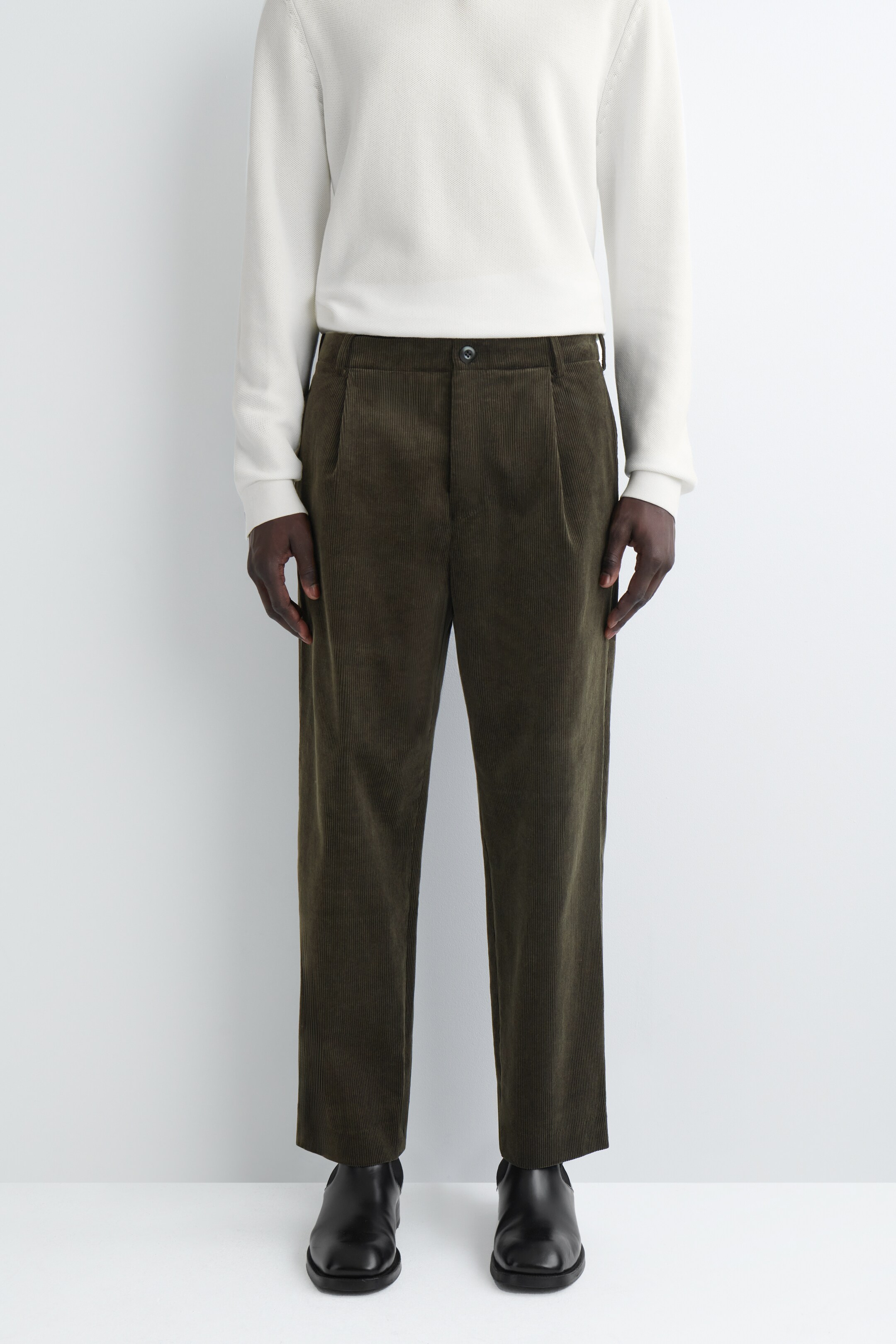 Agrandir l'image: Le pantalon en velours côtelé vert olive foncé est montré de face, avec une coupe décontractée, des plis simples et une longueur légèrement raccourcie. Ils sont portés avec un pull crème et des chaussures noires, mettant en valeur leur silhouette complète et leur ourlet.