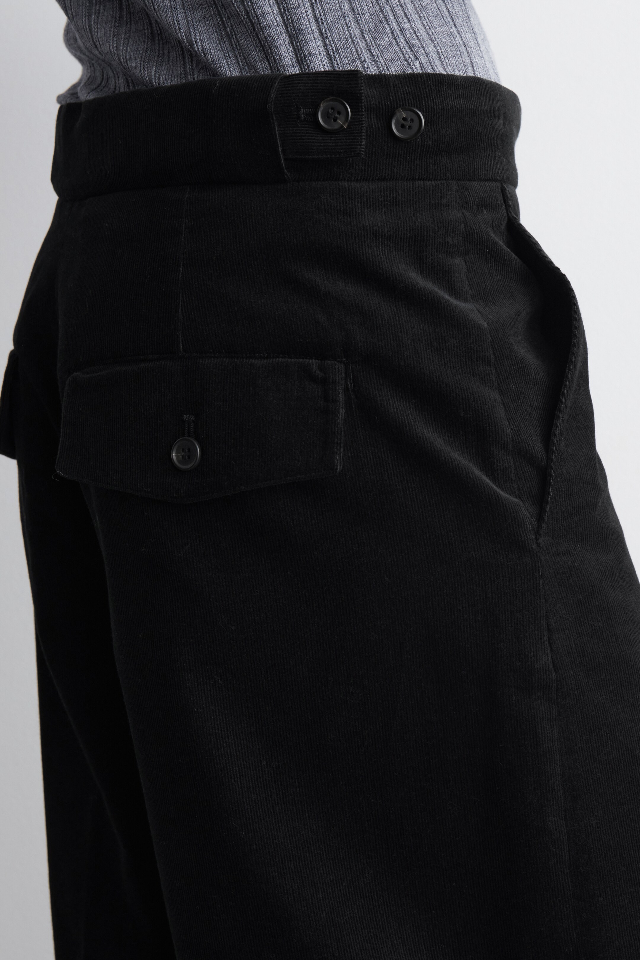 Größeres Bild ansehen: Schwarze Cordhosen werden aus einem seitlichen Winkel präsentiert, um ihre hoch taillierte Passform und den strukturierten Stoff zu zeigen. Sie verfügen über einen breiten Bund mit einem Zwei-Knopf-Verschluss, eine Seitentasche und eine Klappentasche mit Knopfverschluss am linken Bein. Ein graues geripptes Oberteil wird in den Bund gesteckt.