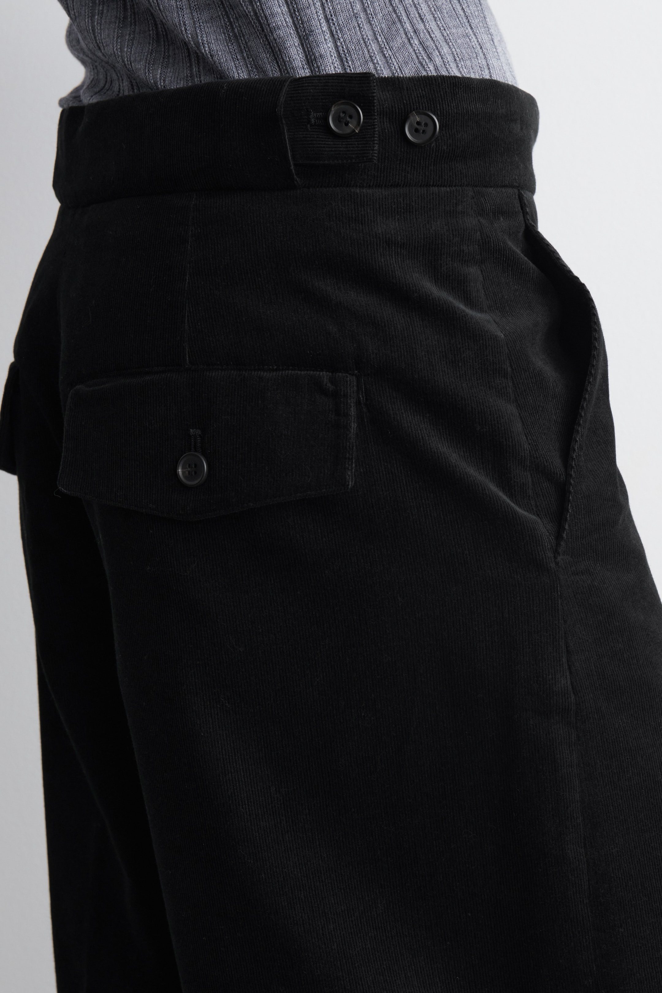 Ver imagen más grande: Se presentan unos pantalones de pana negra desde un ángulo lateral, mostrando su ajuste de cintura alta y su tejido texturizado. Presentan una cinturilla ancha con cierre de lengüeta de dos botones, un bolsillo en la costura lateral y un bolsillo con solapa y cierre de botón en la pierna izquierda. Una camiseta gris de canalé está metida en la cinturilla.