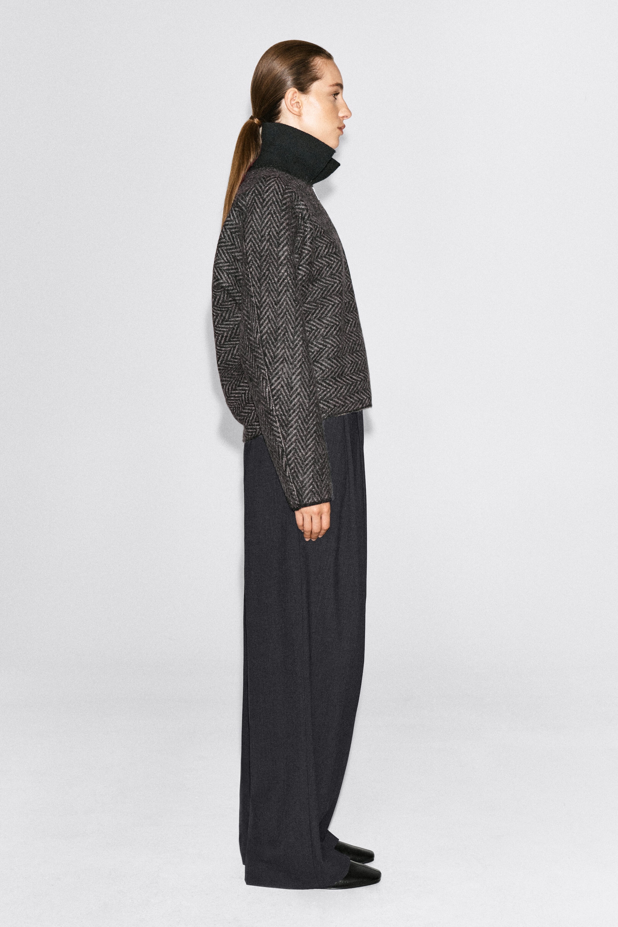 Une veste en tricot à chevrons raccourcie, dotée d’un col côtelé noir montant, est portée par le mannequin, qui se tient de profil. La veste est assortie à un pantalon foncé à jambe large.
