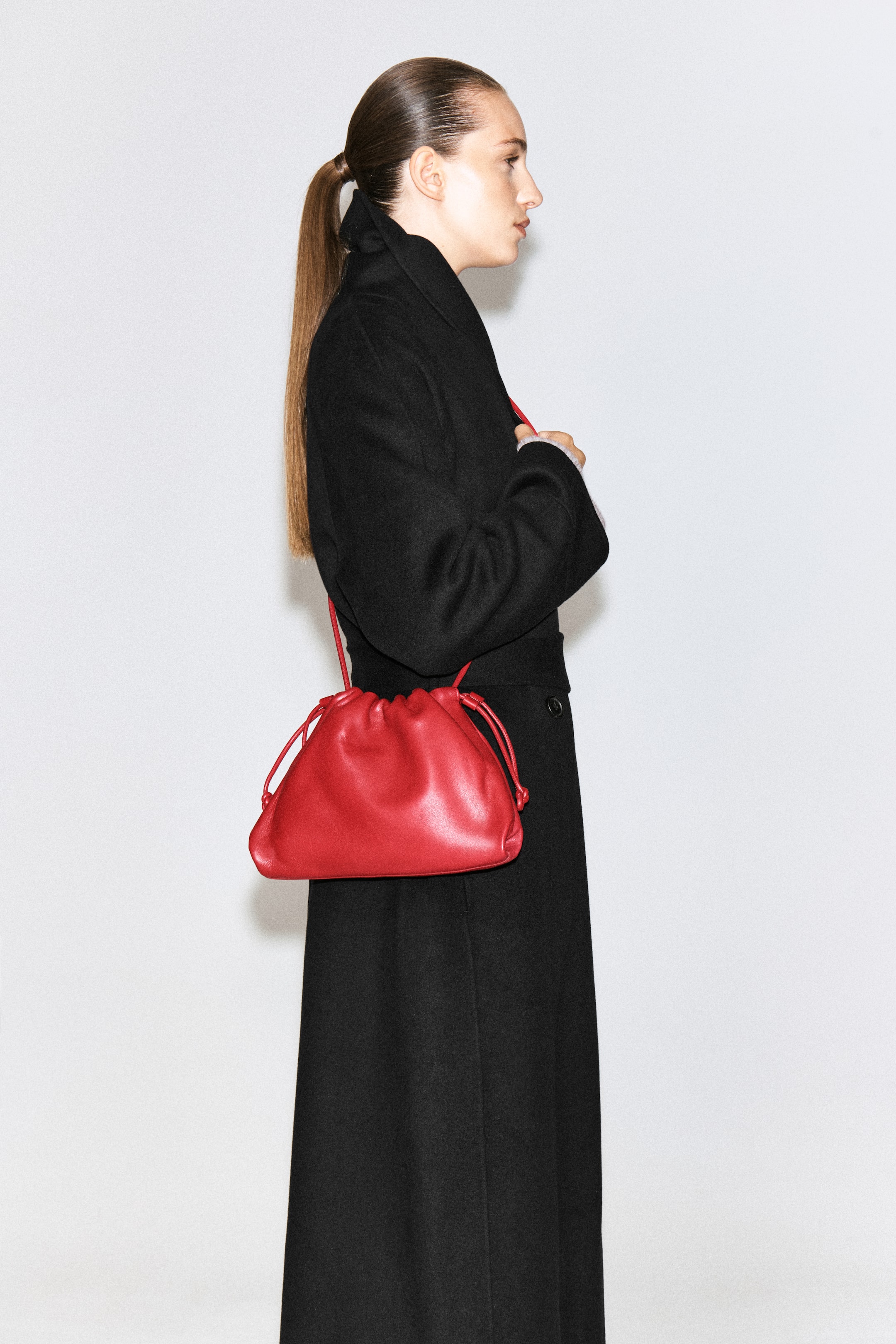 Ver imagen más grande: El bolso clutch Mini Cavatelli de piel roja se lleva al hombro por la modelo, que viste un abrigo largo negro. El bolso presenta una silueta fruncida, tipo bolsa, con finos detalles laterales de cordón ajustable y una correa de hombro a juego de piel.