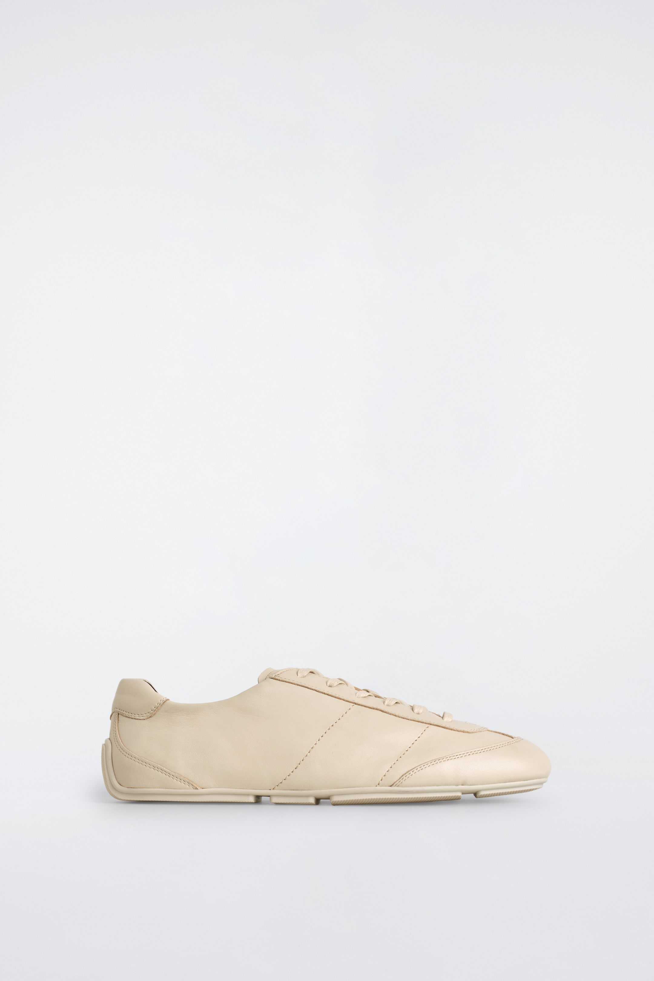 Größeres Bild ansehen: Ein einzelner hellbeiger Ledersneaker ist nach rechts ausgerichtet und präsentiert eine stromlinienförmige Silhouette. Er verfügt über einen tonalen Schnürverschluss, sichtbare Ziernähte, die entlang der Seiten Paneele bilden, und eine vierteilige Gummisohle.