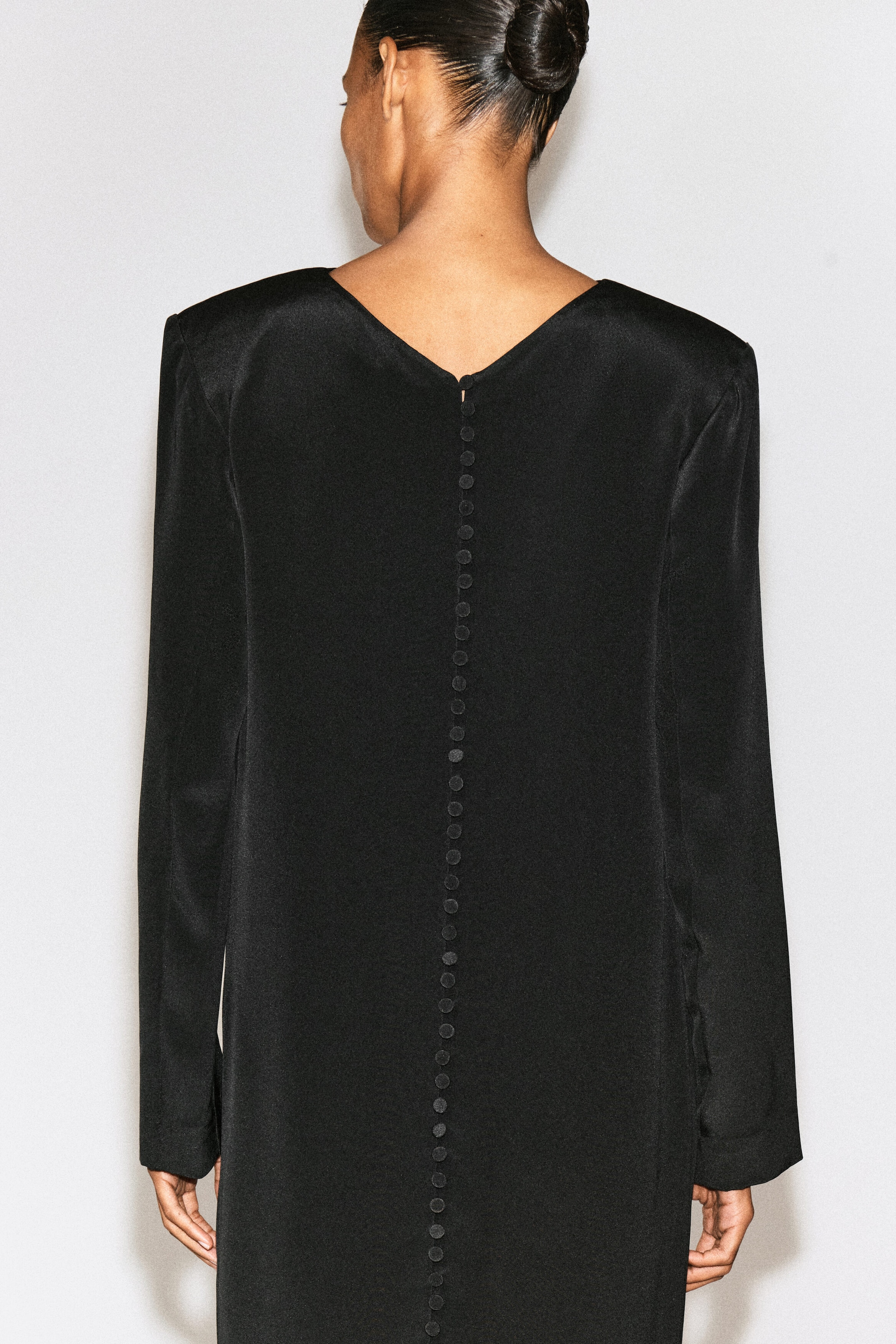 A black, long-sleeved dress with padded shoulders and a V-shaped neckline is worn by the model, shown from behind. A long vertical row of small, fabric-covered buttons runs down the centre back of the dress.