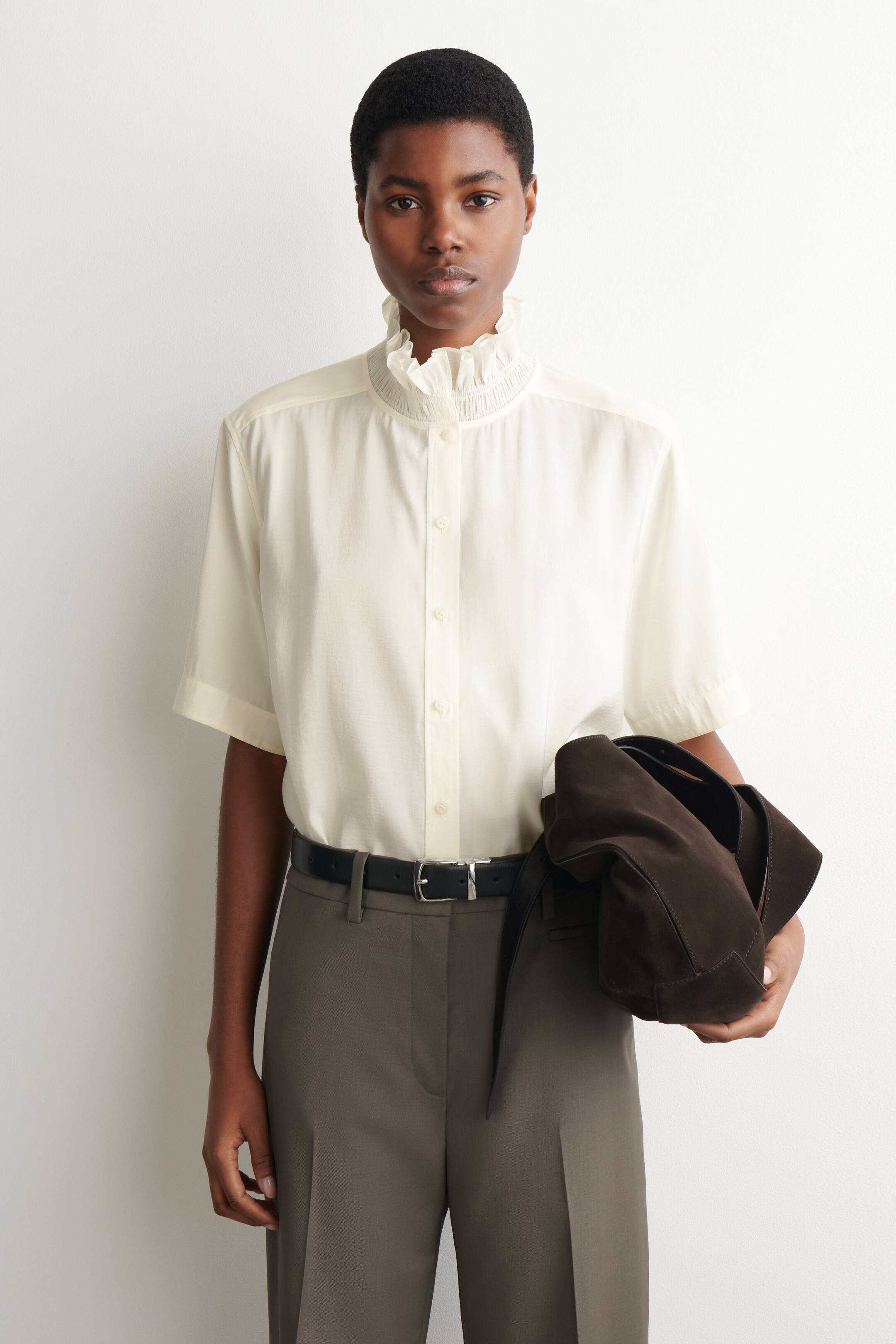 Ver imagen más grande: Una blusa de manga corta en color crema presenta un cuello alto con volantes y detalles de nido de abeja, junto con una tapeta de botones en la parte delantera. Se lleva con pantalones de cintura alta en gris oscuro, asegurados con un cinturón negro con hebilla plateada.