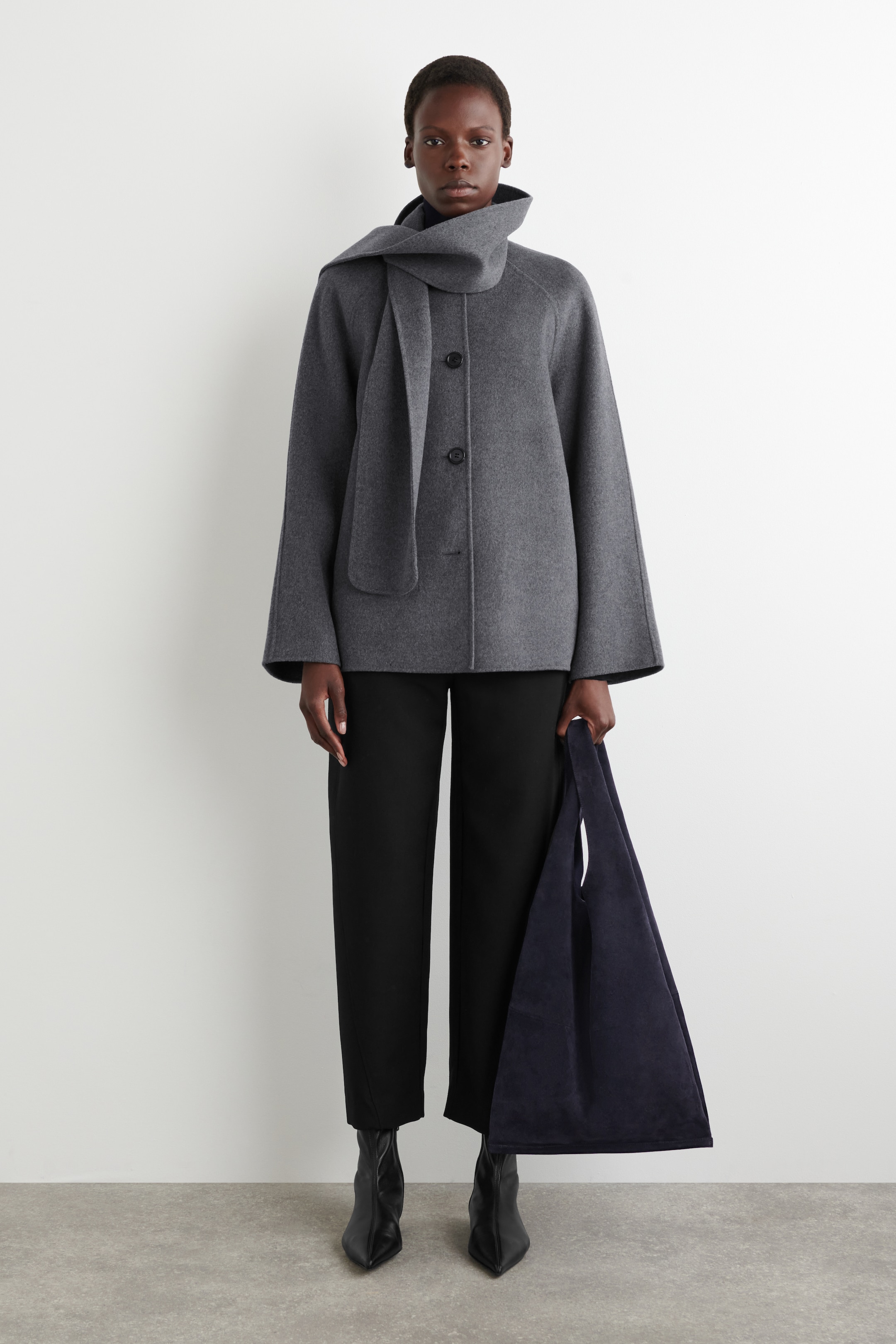 Visualizza immagine più grande: Un cappotto grigio scuro, corto e a vestibilità ampia viene presentato da un'angolazione frontale, caratterizzato da maniche raglan, polsini larghi e tre grandi bottoni frontali. Una sciarpa grigio scuro abbinata è drappeggiata intorno al collo alto del cappotto, che è abbinato a un dolcevita nero.