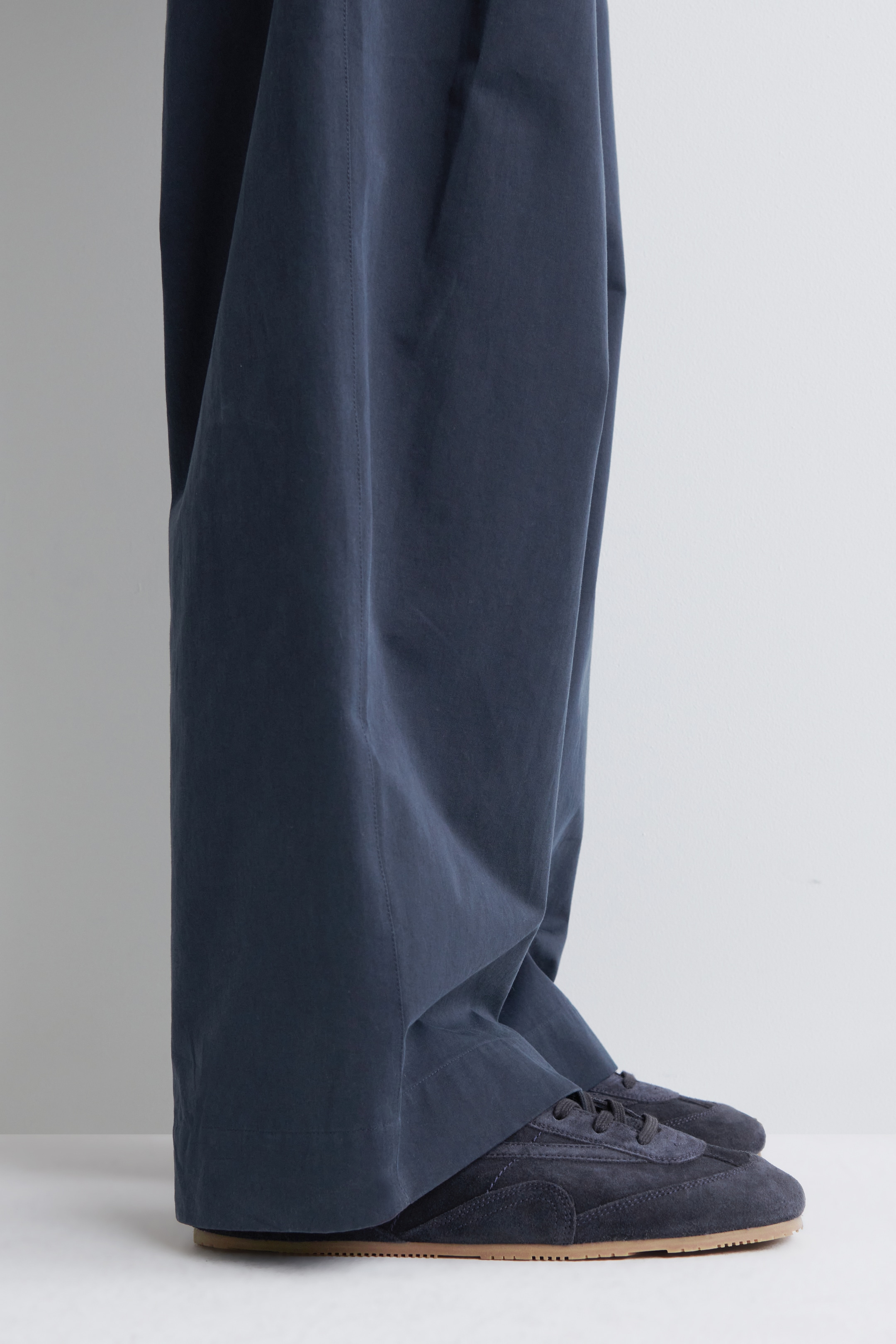 Visualizza immagine più grande: Pantaloni blu scuro a gamba larga si accumulano su un paio di sneakers in camoscio blu scuro abbinate. I pantaloni presentano una cucitura verticale visibile che corre lungo il lato di ciascuna gamba, mentre le sneakers mostrano suole in gomma marrone chiaro.