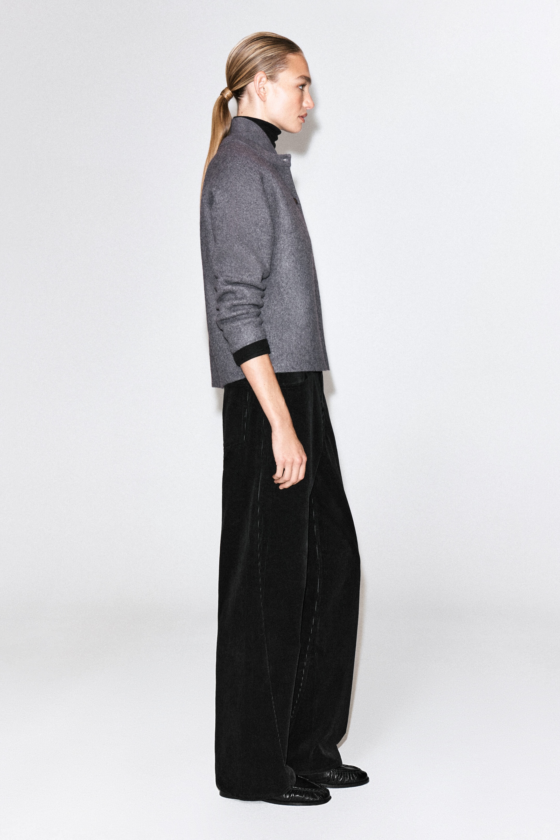 A gray boiled wool cardigan with a rounded silhouette and high collar is worn by the model, who is standing in profile. The cardigan is layered over a black turtleneck top and styled with black wide-leg trousers. A gray boiled wool cardigan with a rounded silhouette and high collar is worn by the model, who is standing in profile. The cardigan is layered over a black turtleneck top and styled with black wide-leg trousers.