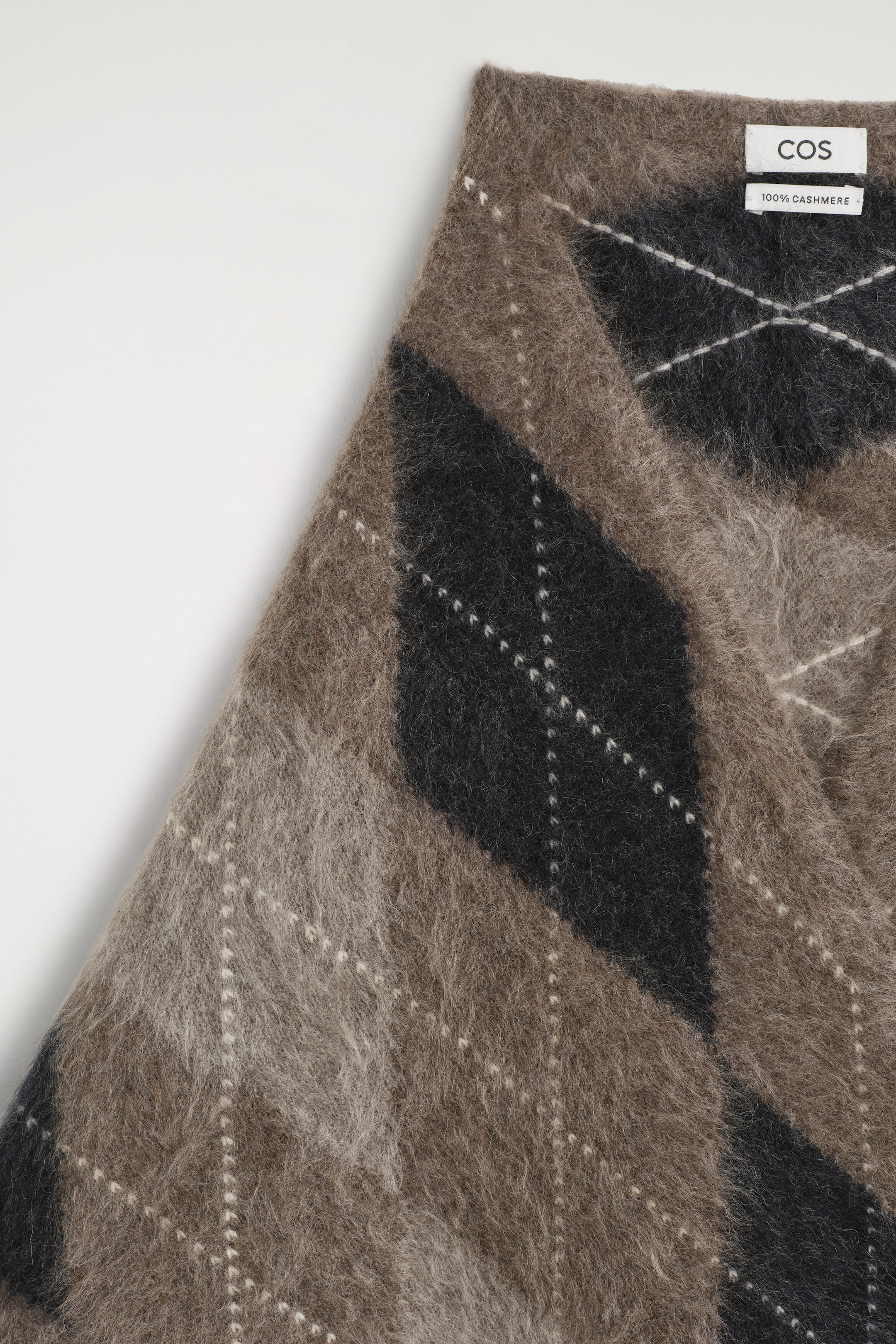 The argyle brushed cashmere triangle scarf showcases a soft, fuzzy texture and a classic argyle pattern in shades of brown, grey, and black with white dashed lines. A white brand label displaying "COS" and "100% CASHMERE" appears on the upper corner of the folded fabric. The argyle brushed cashmere triangle scarf showcases a soft, fuzzy texture and a classic argyle pattern in shades of brown, grey, and black with white dashed lines. A white brand label displaying "COS" and "100% CASHMERE" appears on the upper corner of the folded fabric.