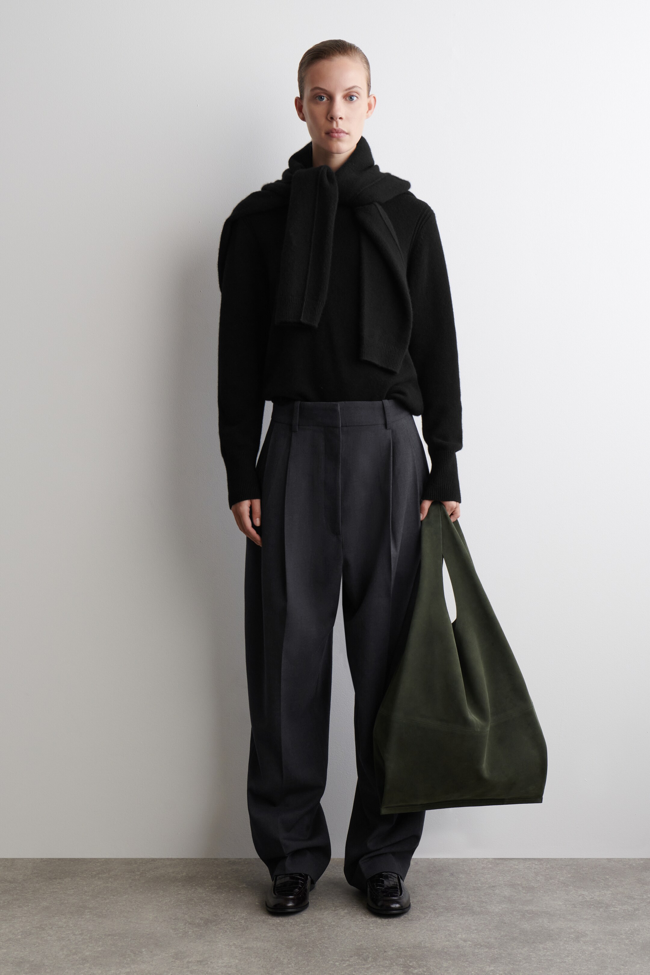 Agrandir l'image: Un pull noir à col rond avec une coupe décontractée est porté, superposé d'une écharpe en tricot noir drapée autour du cou. Un pantalon large plissé gris foncé est porté, associé à des mocassins en cuir verni noir et un sac hobo vert foncé doux tenu à la main.