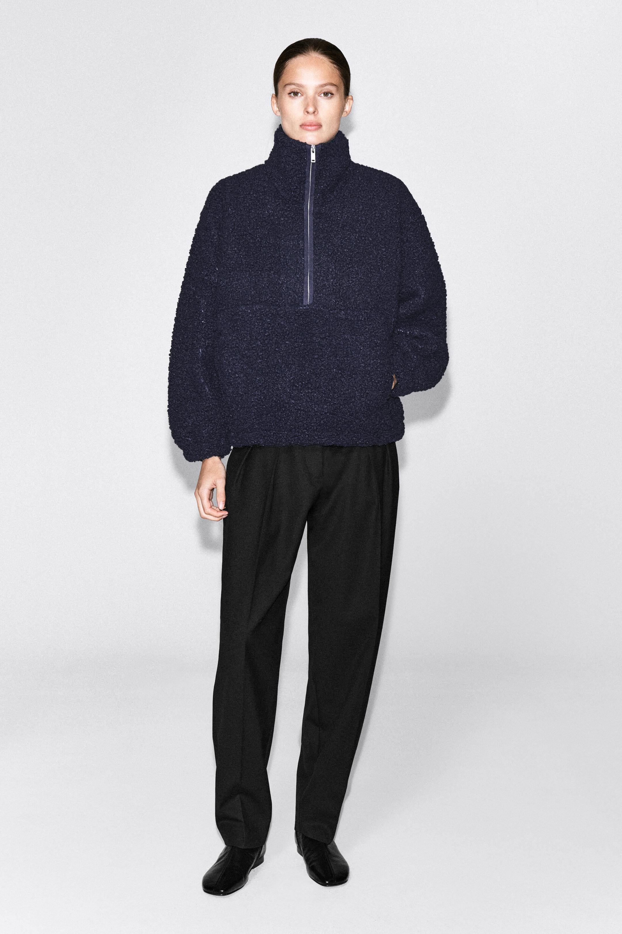The model is wearing a dark navy blue teddy fleece half-zip jumper and standing with one hand in a pocket. The relaxed-fit jumper features a high funnel neck and a silver-toned front zipper, and is styled with black tailored trousers. The model is wearing a dark navy blue teddy fleece half-zip jumper and standing with one hand in a pocket. The relaxed-fit jumper features a high funnel neck and a silver-toned front zipper, and is styled with black tailored trousers.