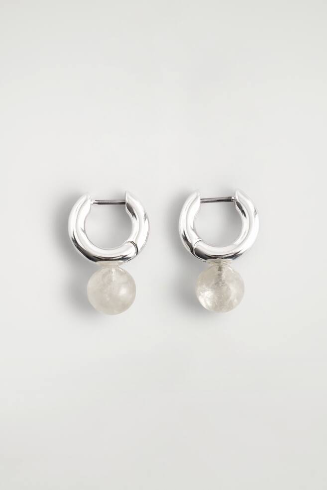 Une paire de boucles d'oreilles présente des anneaux épais de couleur argentée. Chaque anneau suspend une perle sphérique translucide affichant une texture interne nuageuse. - 1