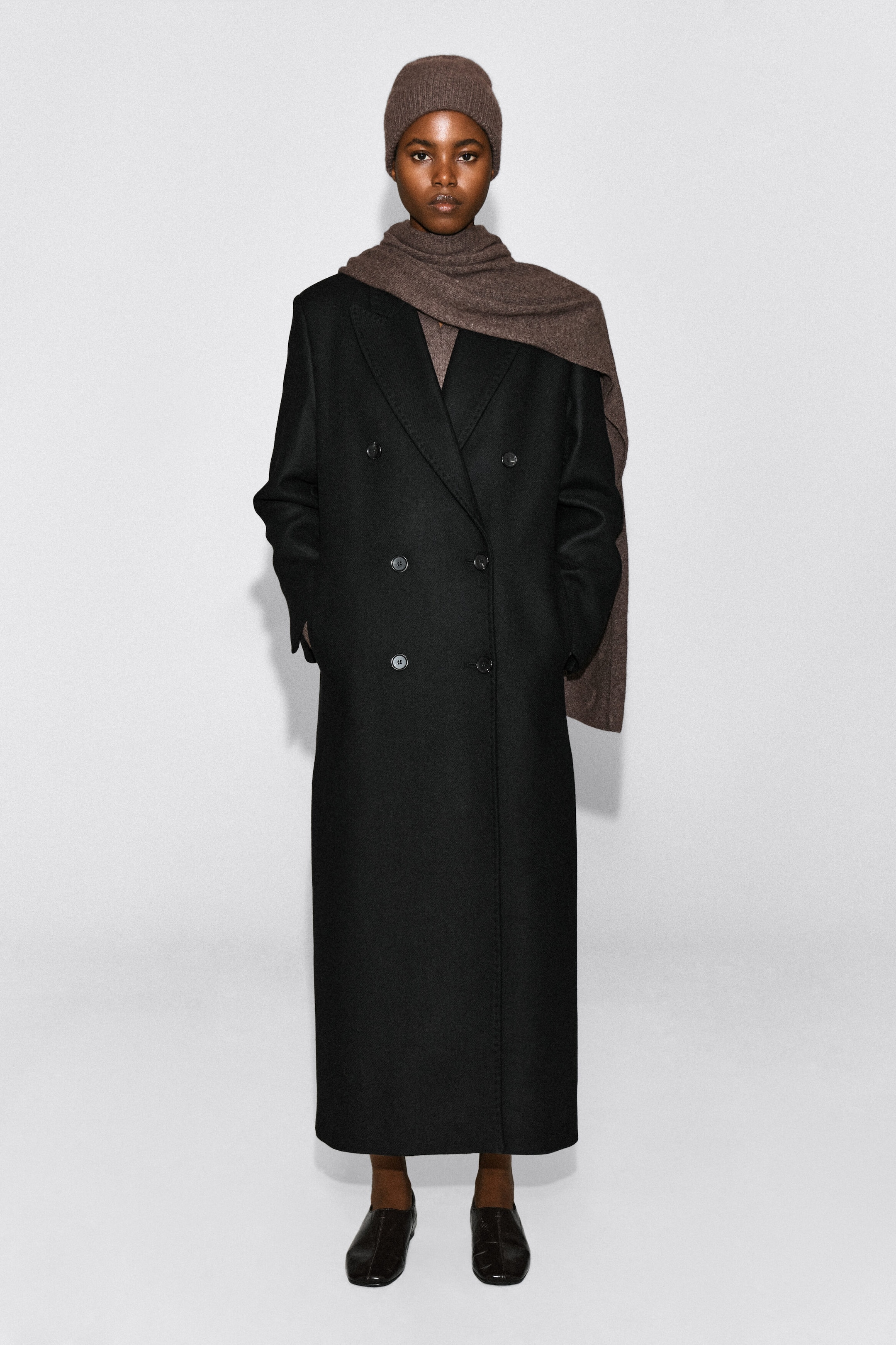 Un bonnet en cachemire brun est porté sur la tête du mannequin, agencé avec une écharpe brune assortie et un long manteau noir à double boutonnage. Le bonnet présente une fine texture en tricot côtelé et épouse bien le front.