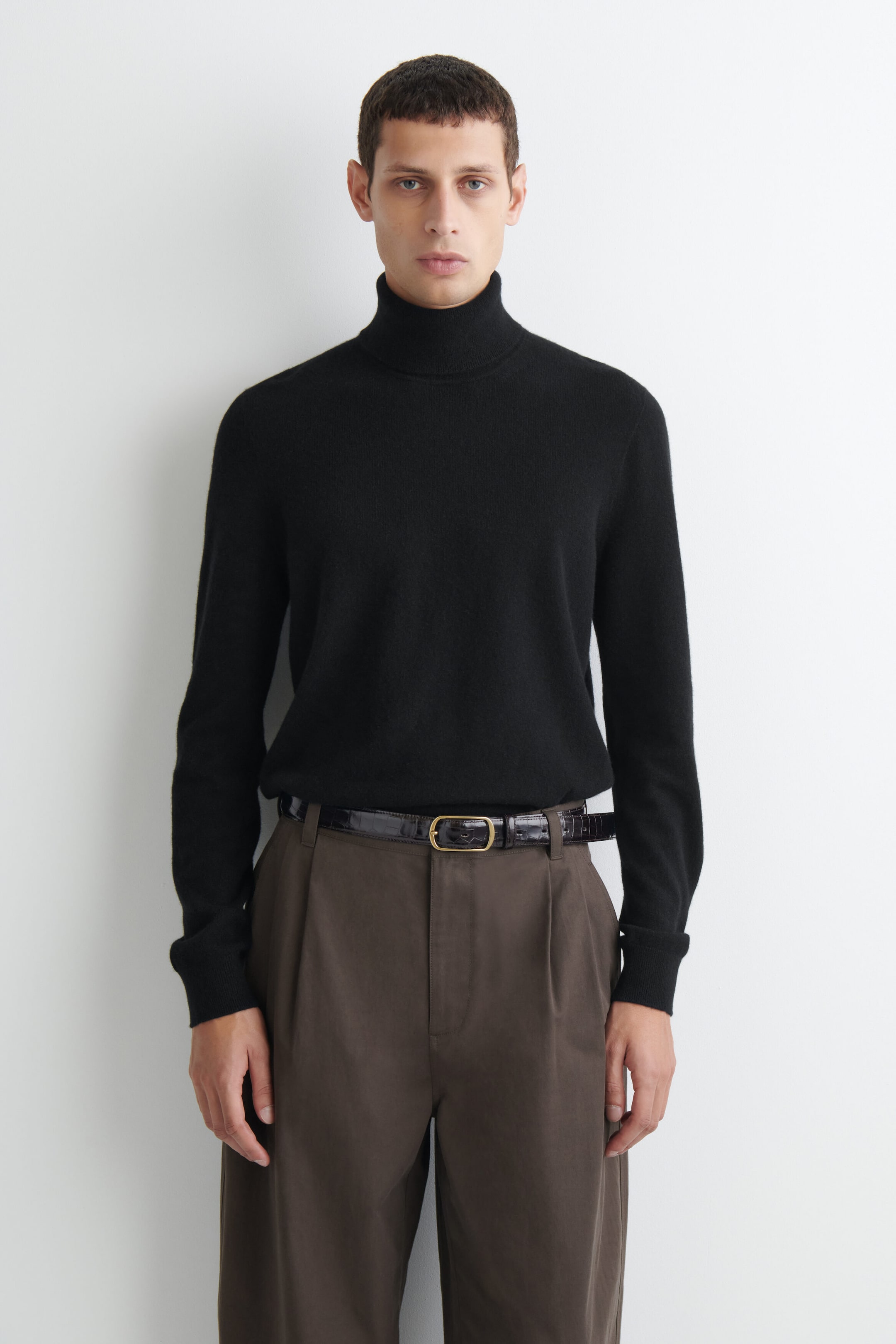 This black turtleneck sweater is shown from the front, featuring a high roll neck and long sleeves with ribbed cuffs. The fine-knit sweater fits snugly and is tucked into brown trousers with a dark textured belt. This black turtleneck sweater is shown from the front, featuring a high roll neck and long sleeves with ribbed cuffs. The fine-knit sweater fits snugly and is tucked into brown trousers with a dark textured belt.