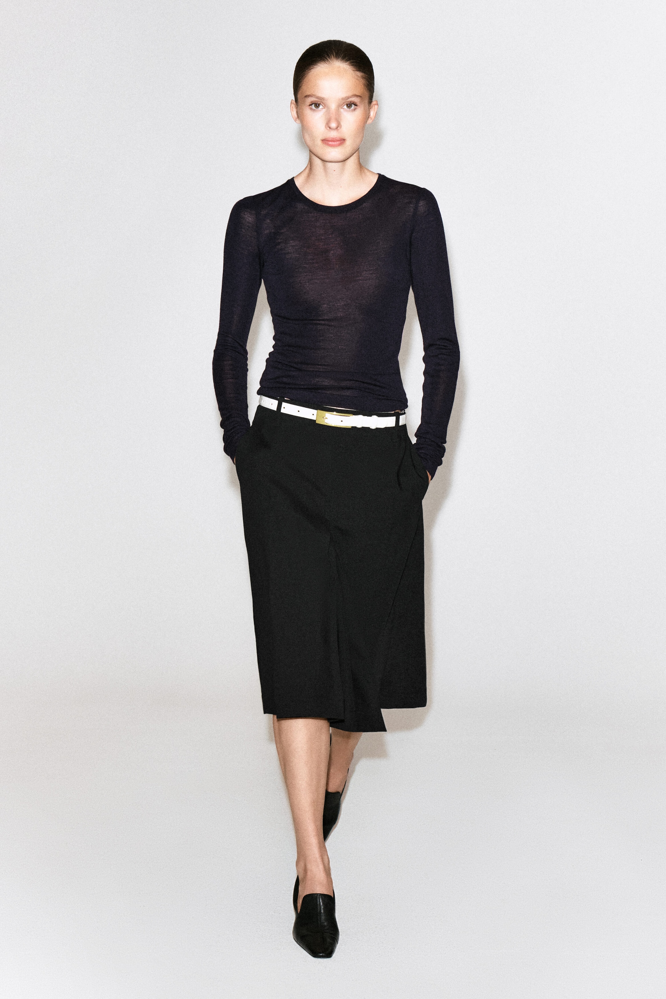 A black box-pleat wool midi skirt is being worn by the model, accessorized with a thin white belt and paired with a sheer, dark long-sleeved top. The skirt features side pockets, into which the model is placing their hands while walking forward. A black box-pleat wool midi skirt is being worn by the model, accessorized with a thin white belt and paired with a sheer, dark long-sleeved top. The skirt features side pockets, into which the model is placing their hands while walking forward.