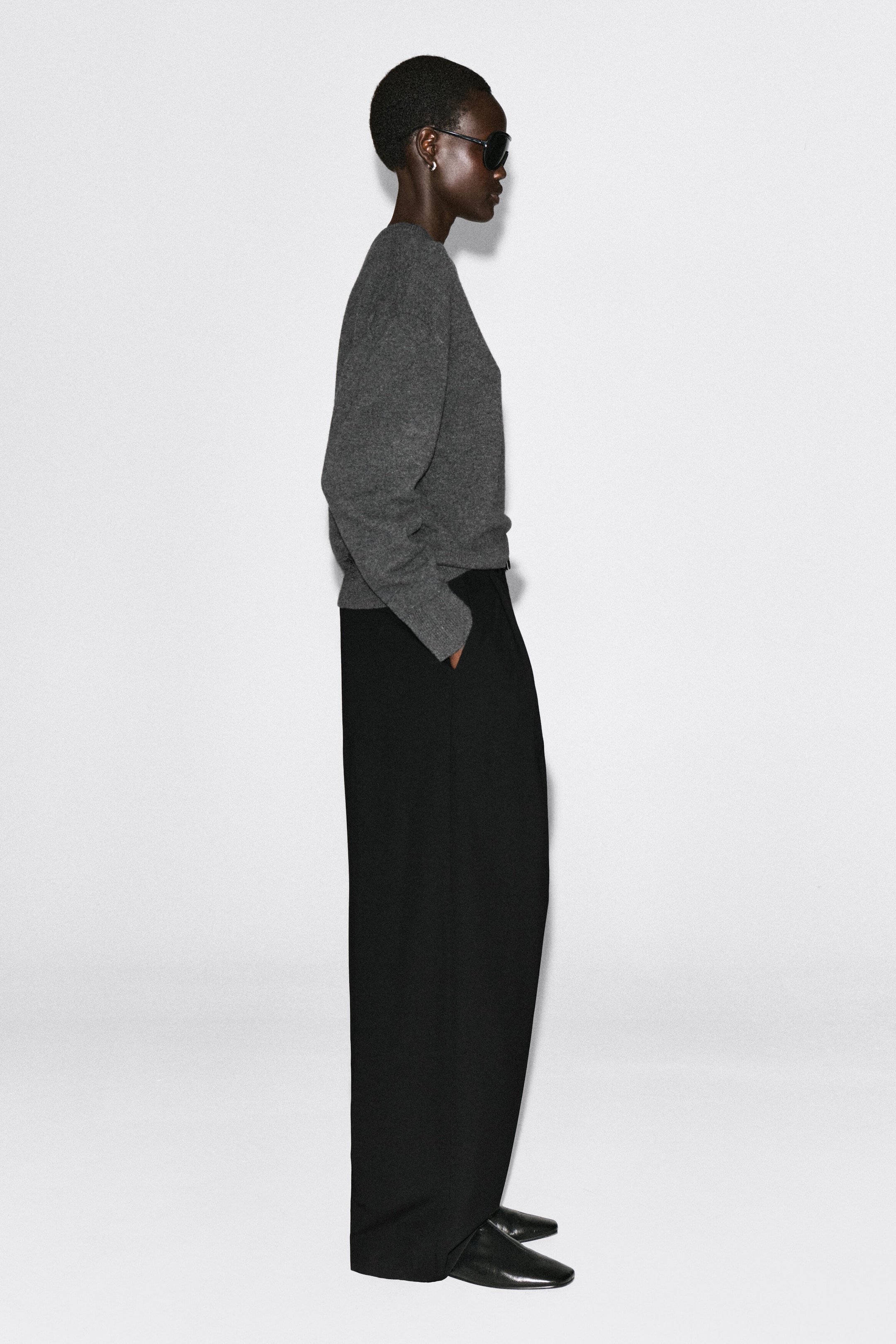 The merino wool layered V-neck jumper is facing right in side profile, showcasing its relaxed fit, dropped shoulders, and ribbed cuffs in a grey fine-knit fabric. The jumper drapes loosely at the waist over black wide-leg trousers to highlight its casual silhouette. The merino wool layered V-neck jumper is facing right in side profile, showcasing its relaxed fit, dropped shoulders, and ribbed cuffs in a grey fine-knit fabric. The jumper drapes loosely at the waist over black wide-leg trousers to highlight its casual silhouette.