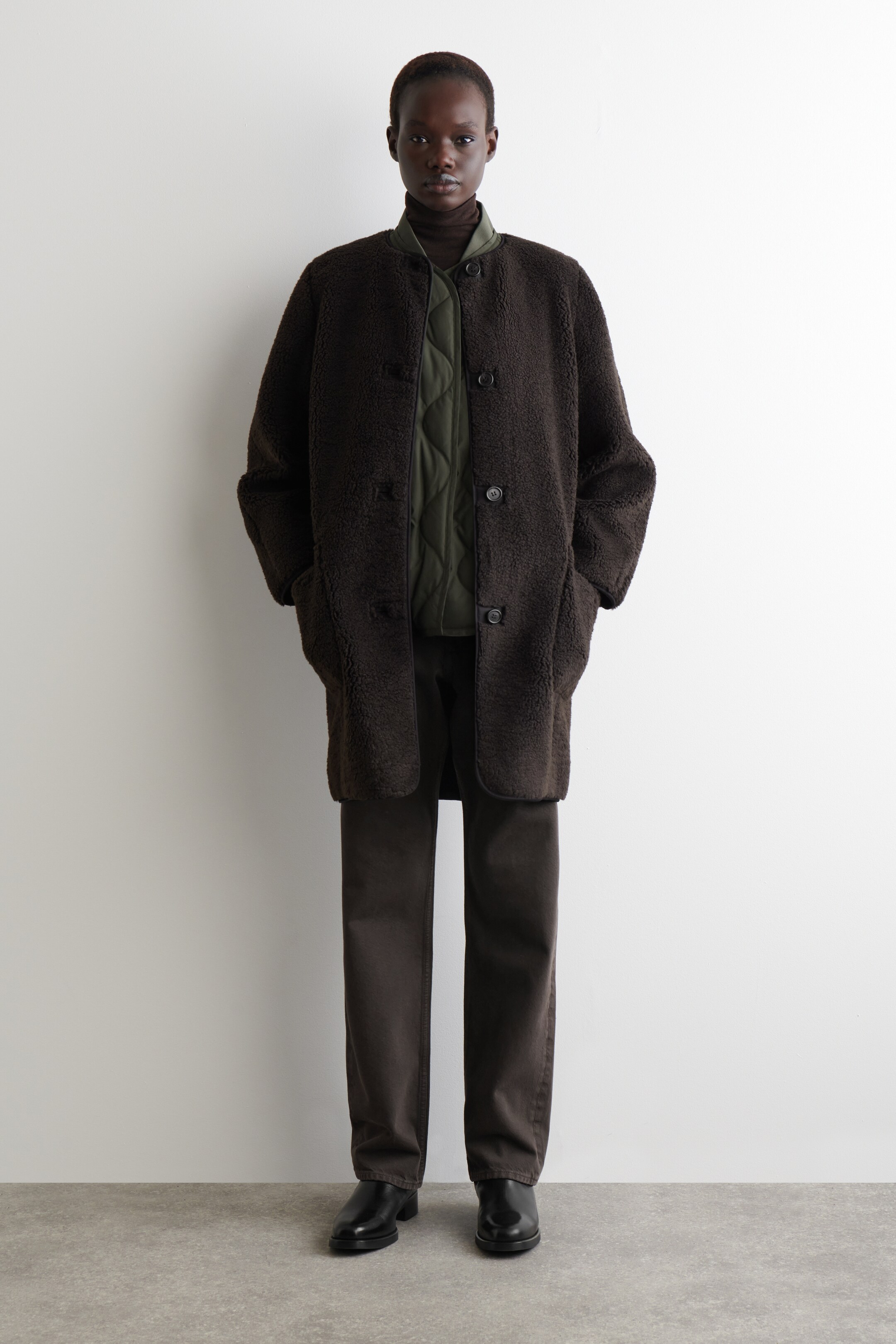 Agrandir l'image: Un manteau en polaire teddy marron foncé est porté face avant, s'étendant jusqu'à mi-cuisse et présentant un design sans col, de gros boutons sombres sur le devant et deux grandes poches avant dans lesquelles les mains sont placées. Un gilet matelassé vert est visible en dessous.