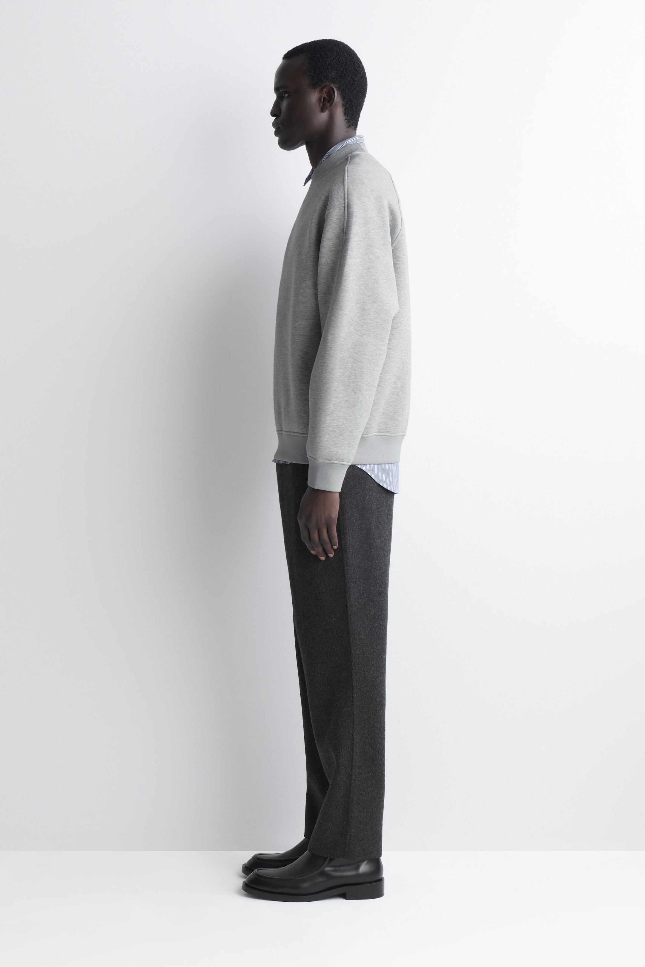 The relaxed scuba sweatshirt in light grey is shown in a relaxed fit from a left-facing side profile, featuring dropped shoulders, ribbed cuffs, and a ribbed hem. It is layered over a striped collared shirt that peeks out at the crew neckline and hem. The relaxed scuba sweatshirt in light grey is shown in a relaxed fit from a left-facing side profile, featuring dropped shoulders, ribbed cuffs, and a ribbed hem. It is layered over a striped collared shirt that peeks out at the crew neckline and hem.
