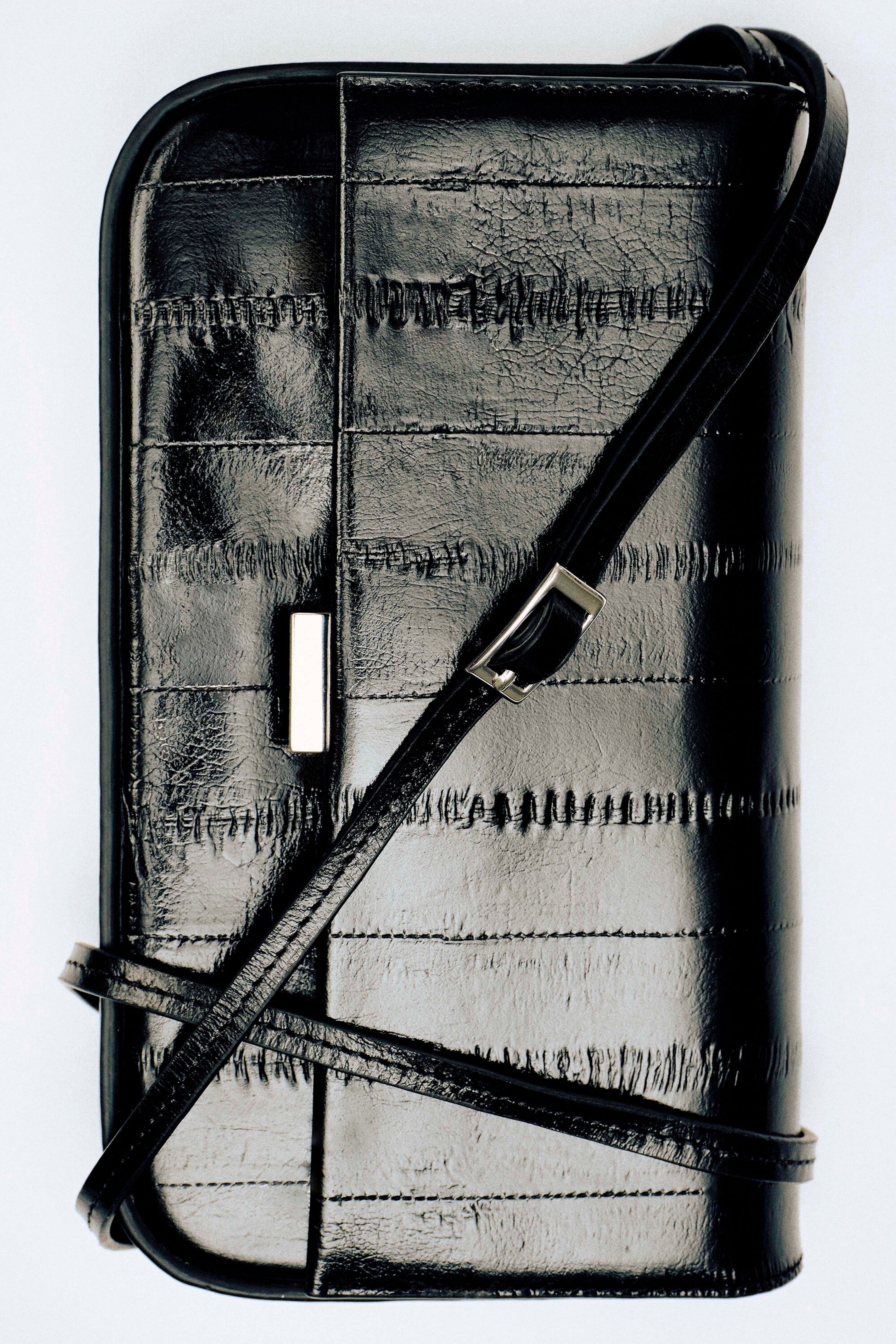 Le sac en cuir noir est présenté de face, mettant en valeur une silhouette rectangulaire pliée, réalisée en cuir à finition brillante avec un motif gaufré à effet anguille, en panneaux. Une fine bandoulière ajustable est drapée en diagonale sur l’avant, ponctuée d’un petit fermoir métallique argenté.