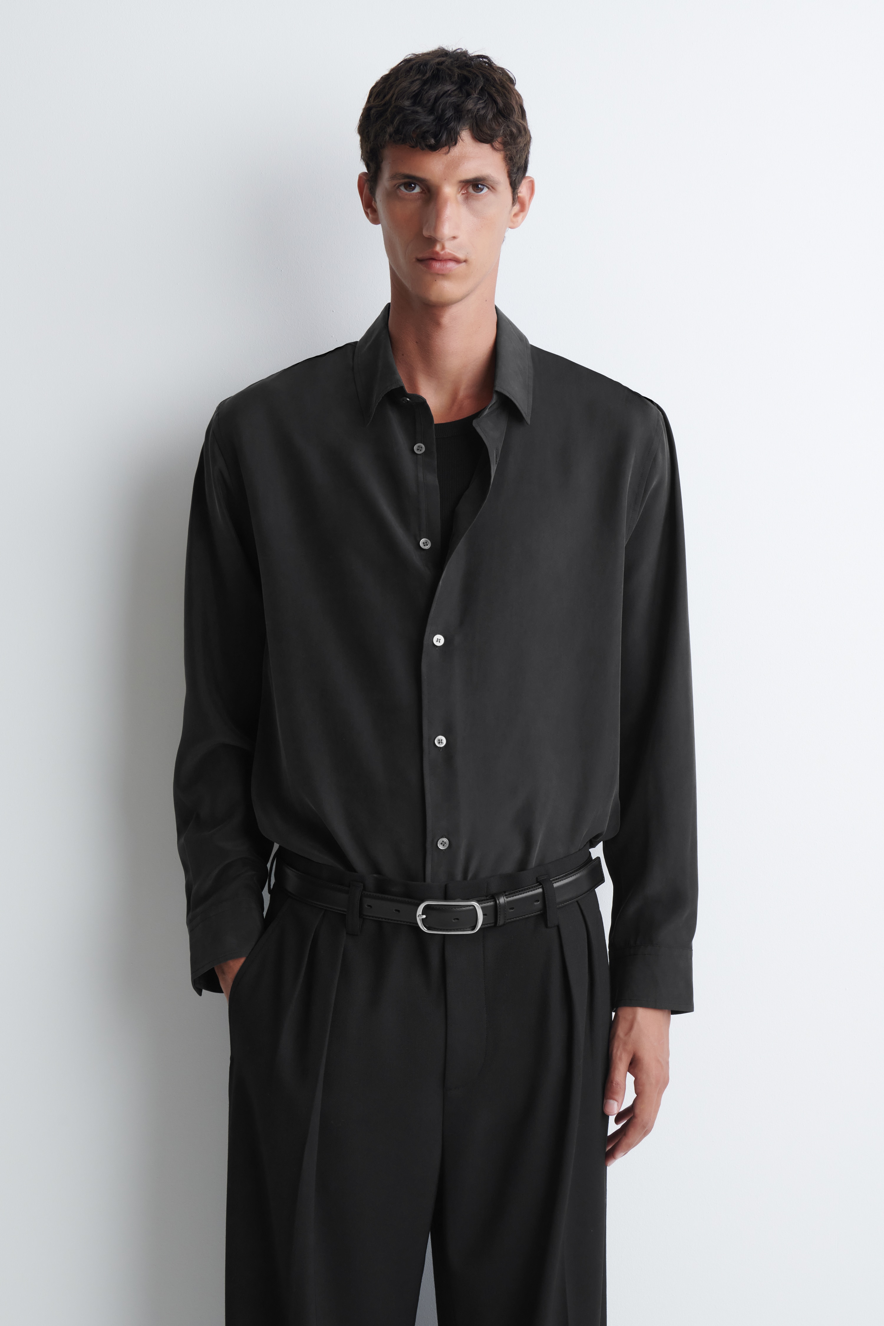 Una camicia in seta sandwashed grigio scuro Ã¨ mostrata frontalmente, con classico colletto a punta, chiusura frontale con bottoni in contrasto di colore chiaro e maniche lunghe. La camicia presenta una vestibilitÃ  rilassata, una texture morbida e opaca e un orlo leggermente arrotondato.