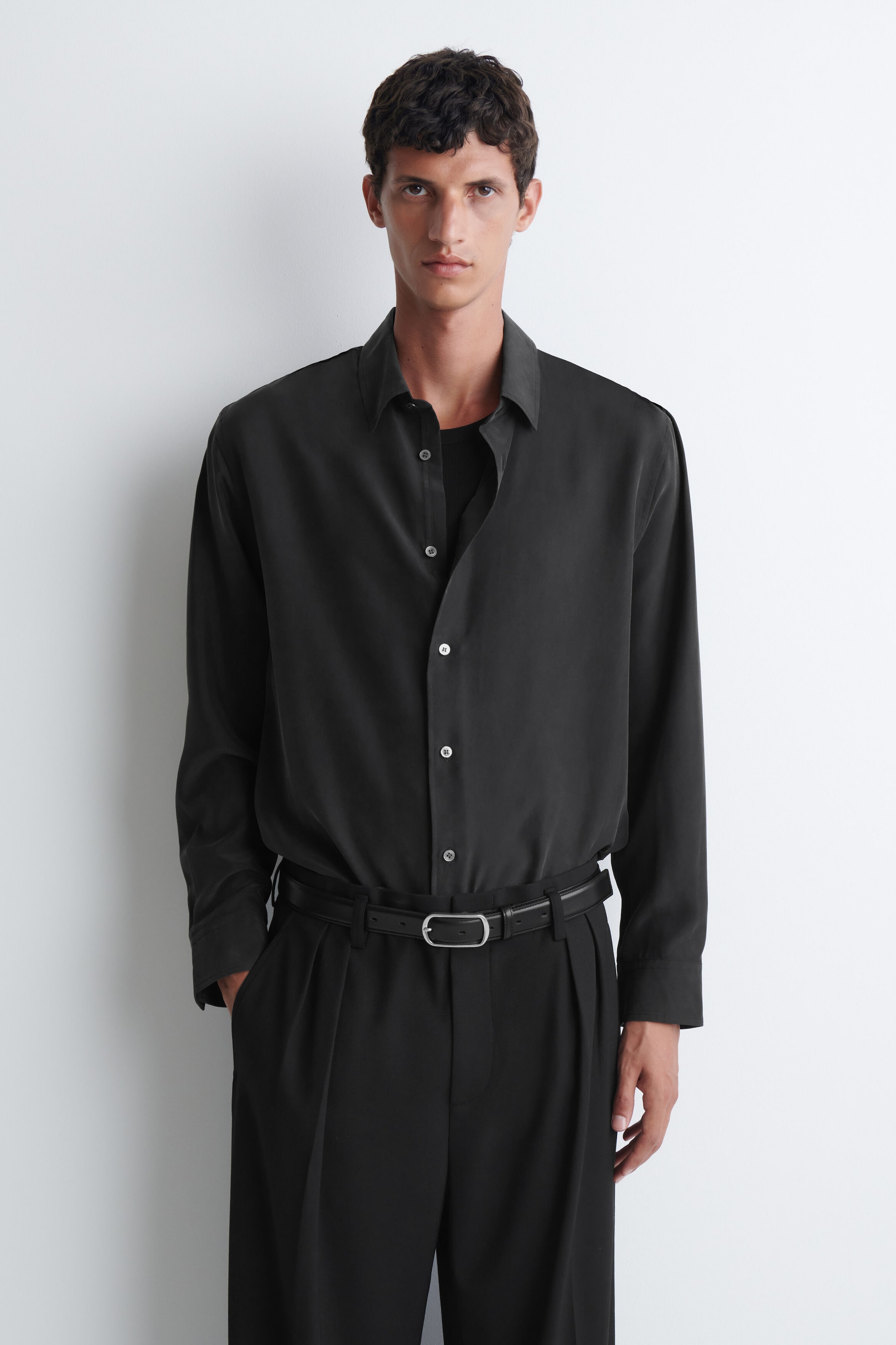 The model is wearing a black sandwashed silk shirt featuring a classic collar, long sleeves, and a button-front fastening. The shirt is styled partially unbuttoned at the collar, layered over a dark top, and tucked into black pleated trousers. The model is wearing a black sandwashed silk shirt featuring a classic collar, long sleeves, and a button-front fastening. The shirt is styled partially unbuttoned at the collar, layered over a dark top, and tucked into black pleated trousers.