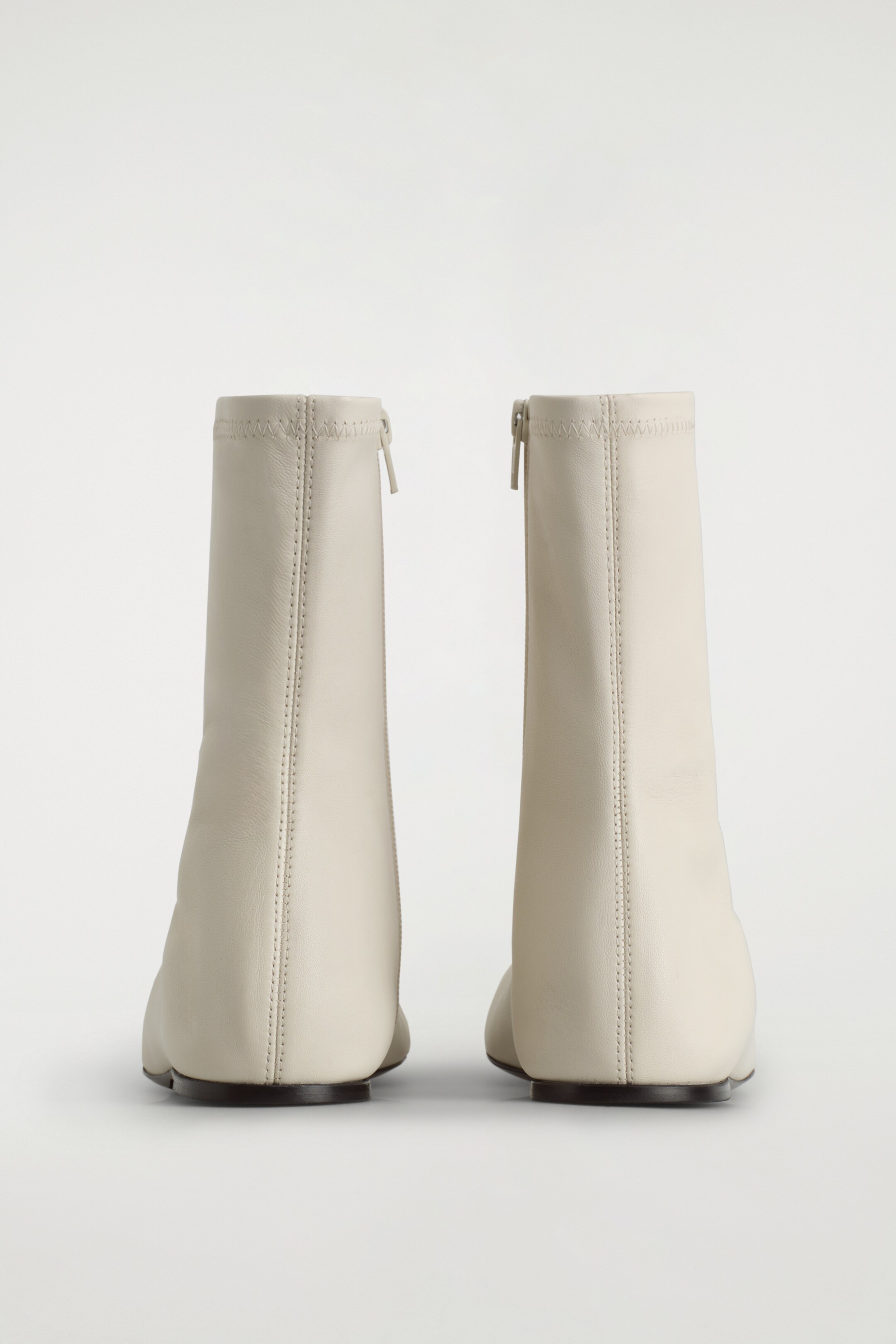 View larger image: Two chalk-toned leather sock boots are standing, viewed from the back, resting on low heels. Each boot features a vertical seam running down the center of the shaft and a metal zip closure extending from the top edge downwards along the back.