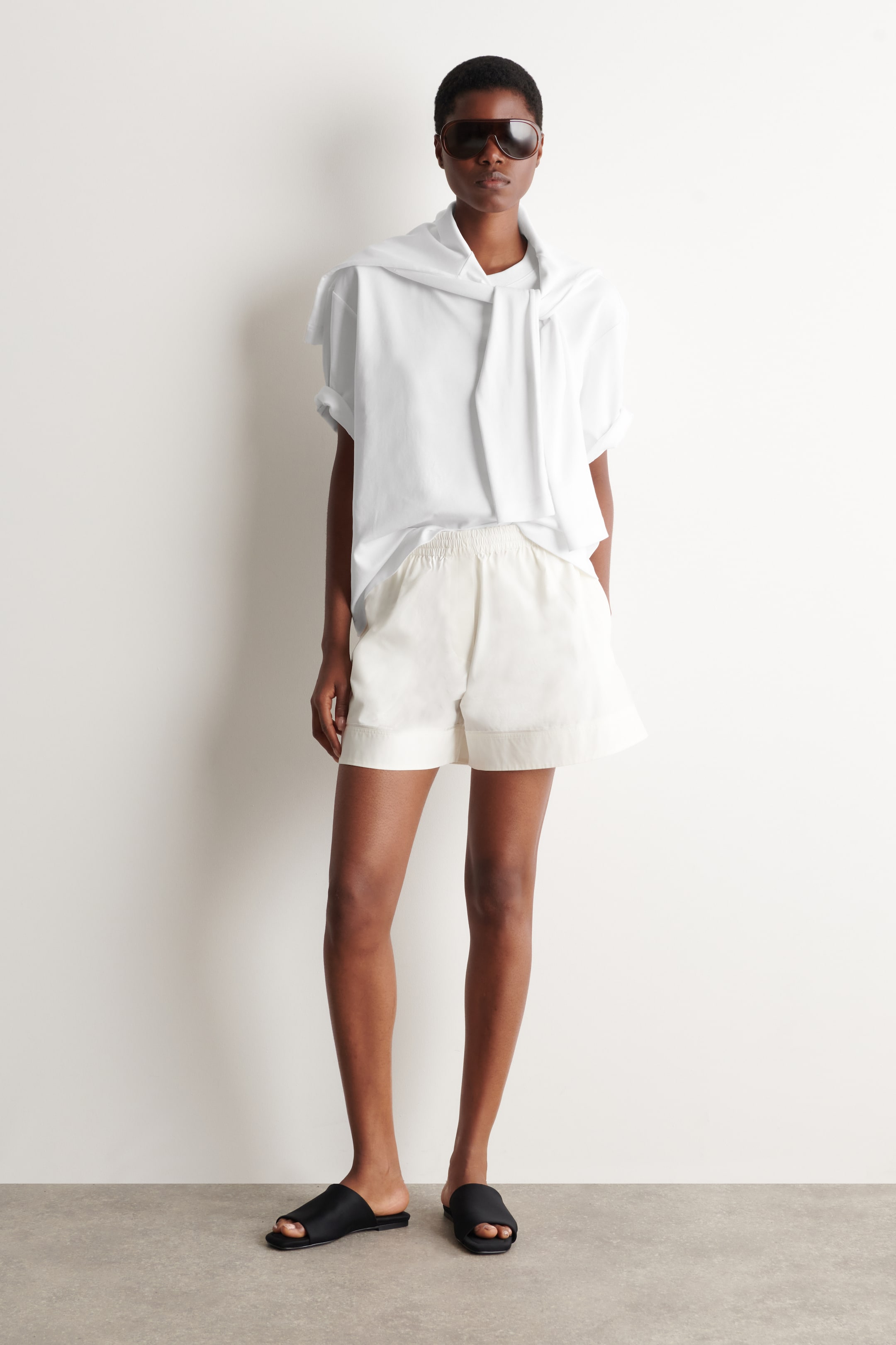 Ver imagem maior: Uma t-shirt branca de manga curta e decote redondo está a ser usada com uma camisa branca de manga comprida, drapeada e amarrada ao redor do pescoço. Uns calções em tom off-white, com cintura elástica e bainhas largas, estão a ser usados com sandálias rasas pretas e óculos de sol grandes e escuros.