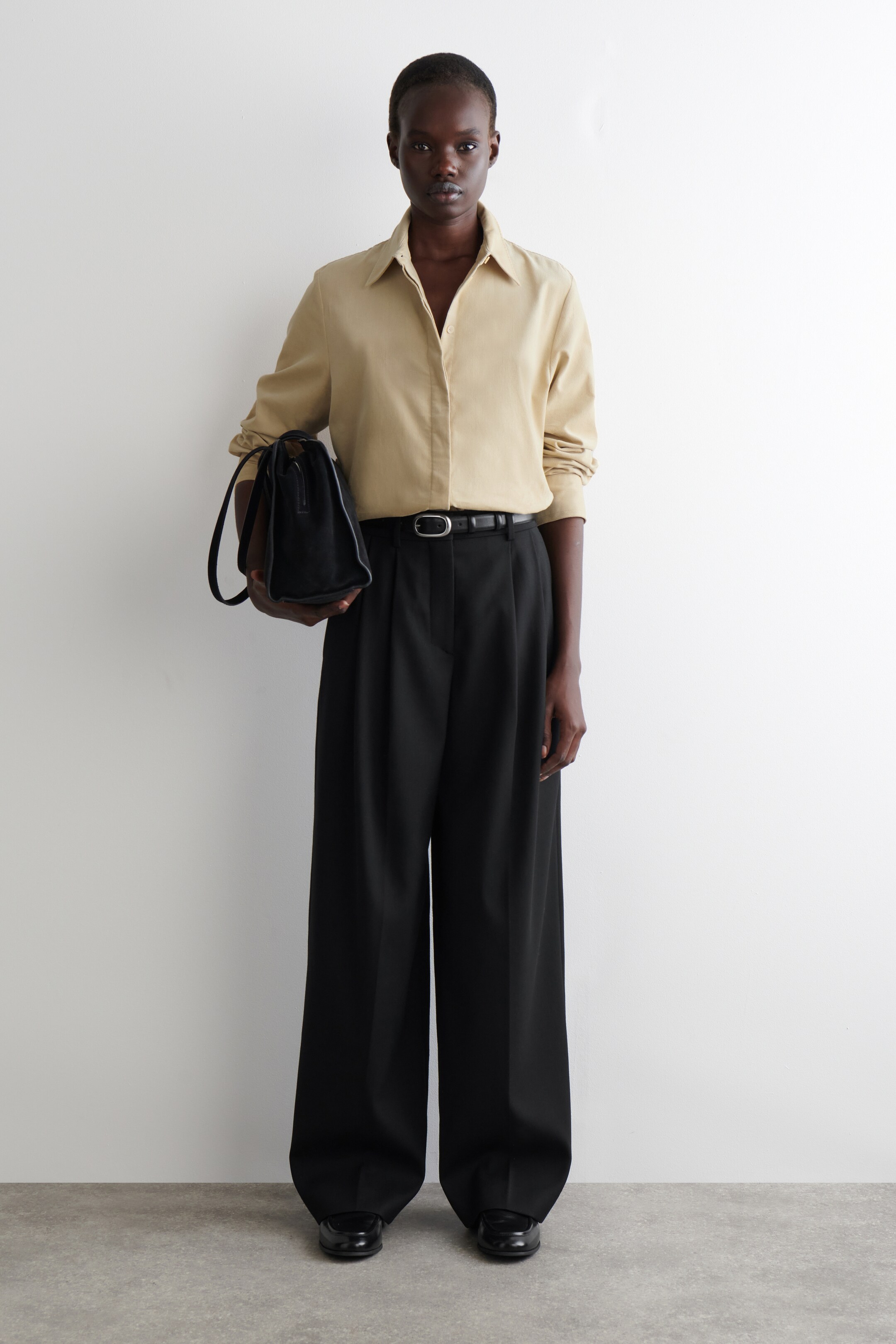 Agrandir l'image: Une chemise boutonnée à manches longues de couleur beige clair avec un col évasé est portée, rentrée dans un pantalon noir à jambes larges et plis, avec une ceinture noire et une boucle ovale en argent. Un sac noir à poignée supérieure est tenu, et des chaussures noires à bout fermé sont visibles, présentant une vue de face de l'ensemble.