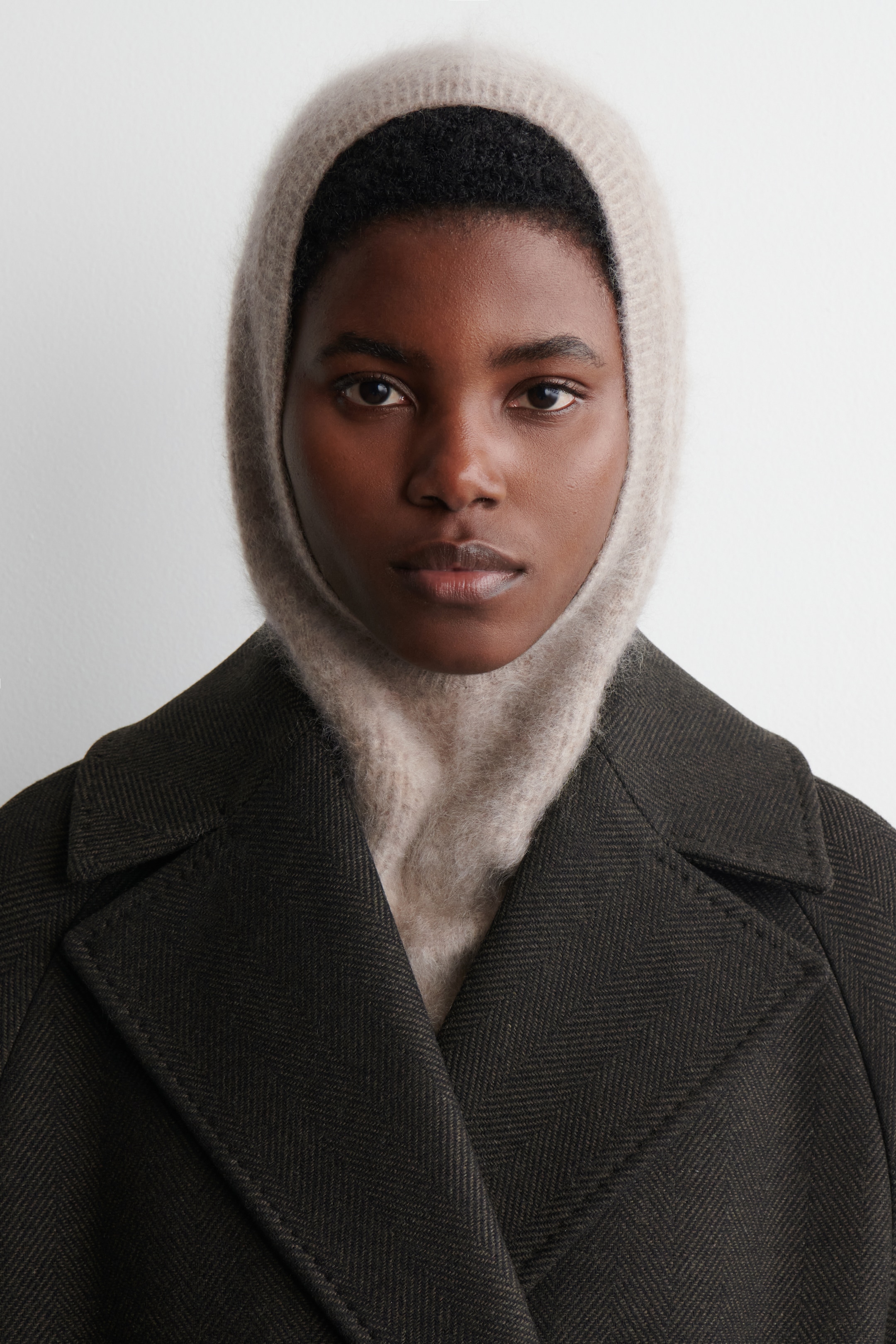 Agrandir l'image: Une cagoule en tricot duveteux beige clair enveloppe la tête et le cou, son ouverture encadrant le visage, et est visible sous les revers crantés structurés d'un manteau vert foncé affichant un tissage en chevrons.