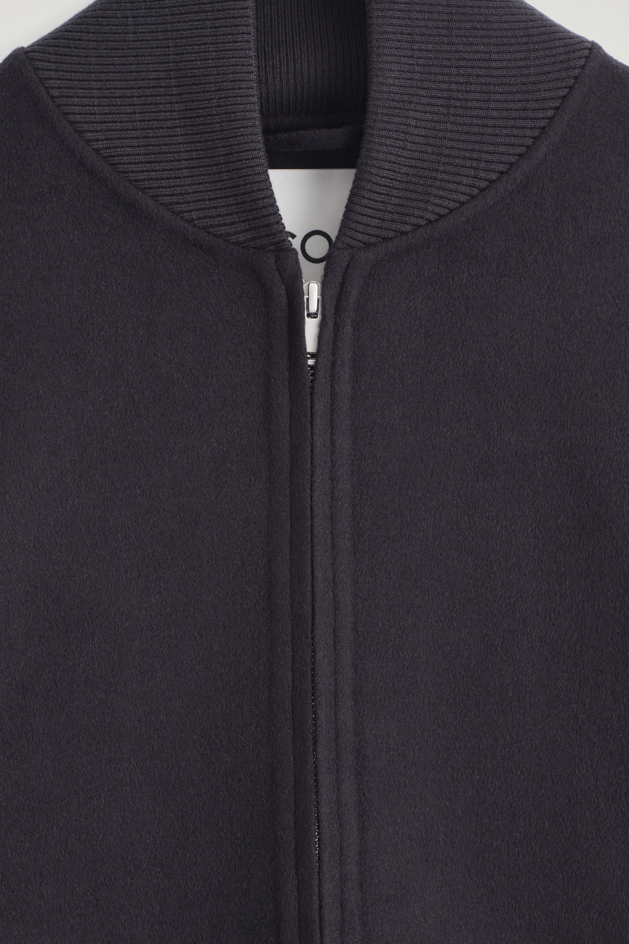 View larger image: A dark grey jacket is being shown from the front, featuring a ribbed knit collar and a silver-toned zipper closure running down its center. A white brand label is appearing at the neckline behind the partially open zipper.