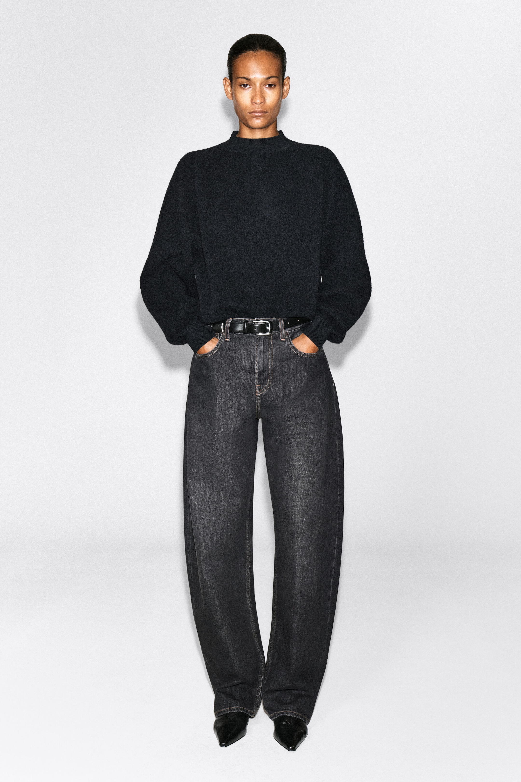The model is wearing dark gray washed wide-leg jeans and posing with her hands in the front pockets. The jeans are styled with a tucked-in black mock neck sweater, a black leather belt with a silver buckle, and black pointed-toe boots. The model is wearing dark gray washed wide-leg jeans and posing with her hands in the front pockets. The jeans are styled with a tucked-in black mock neck sweater, a black leather belt with a silver buckle, and black pointed-toe boots.