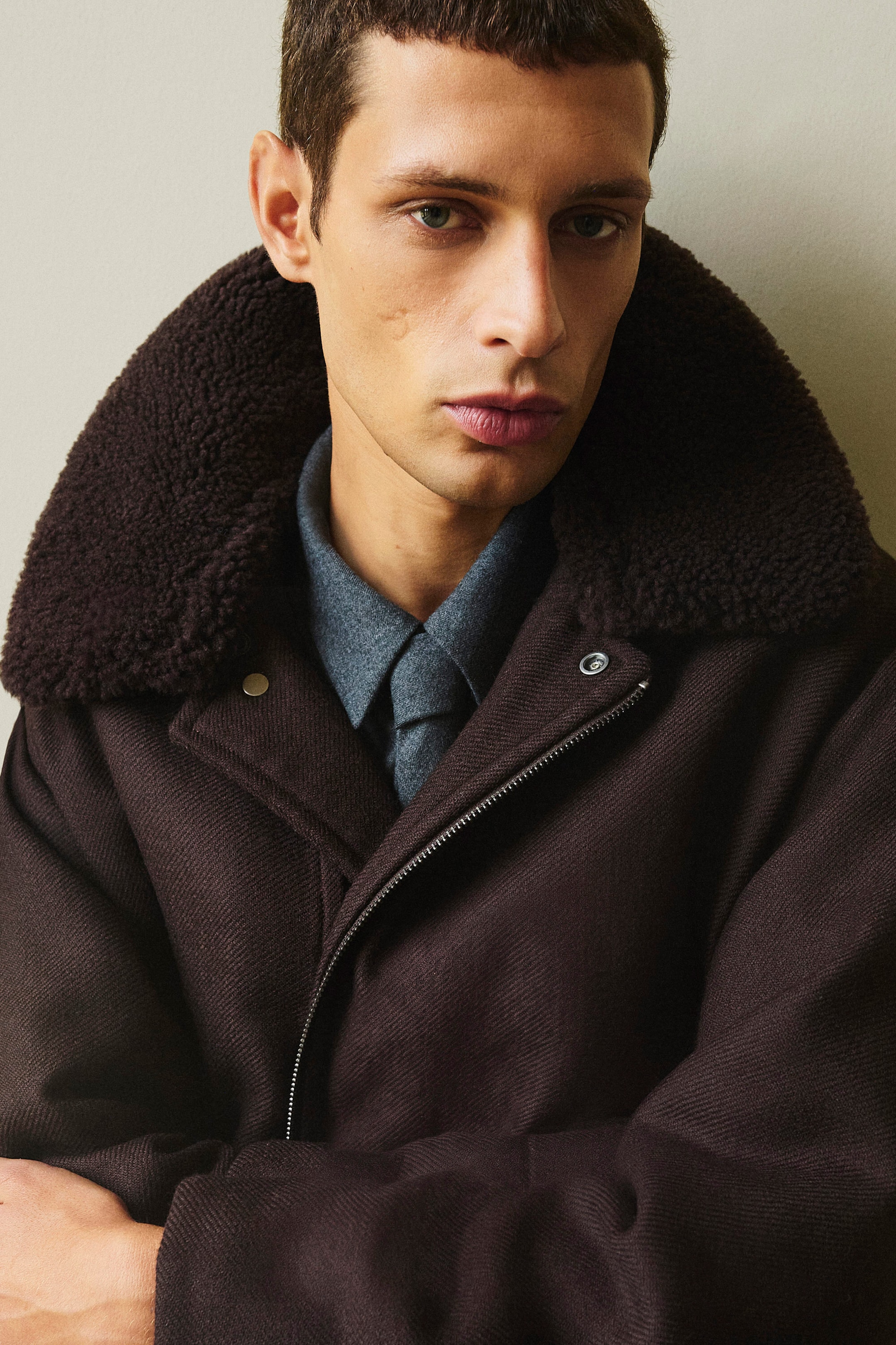 A dark brown wool twill flight jacket is shown from the front, featuring a plush oversized shearling collar and a partially unzipped diagonal fastening. The jacket is layered over a grey wool flannel shirt with a buttoned pointed collar. A dark brown wool twill flight jacket is shown from the front, featuring a plush oversized shearling collar and a partially unzipped diagonal fastening. The jacket is layered over a grey wool flannel shirt with a buttoned pointed collar.