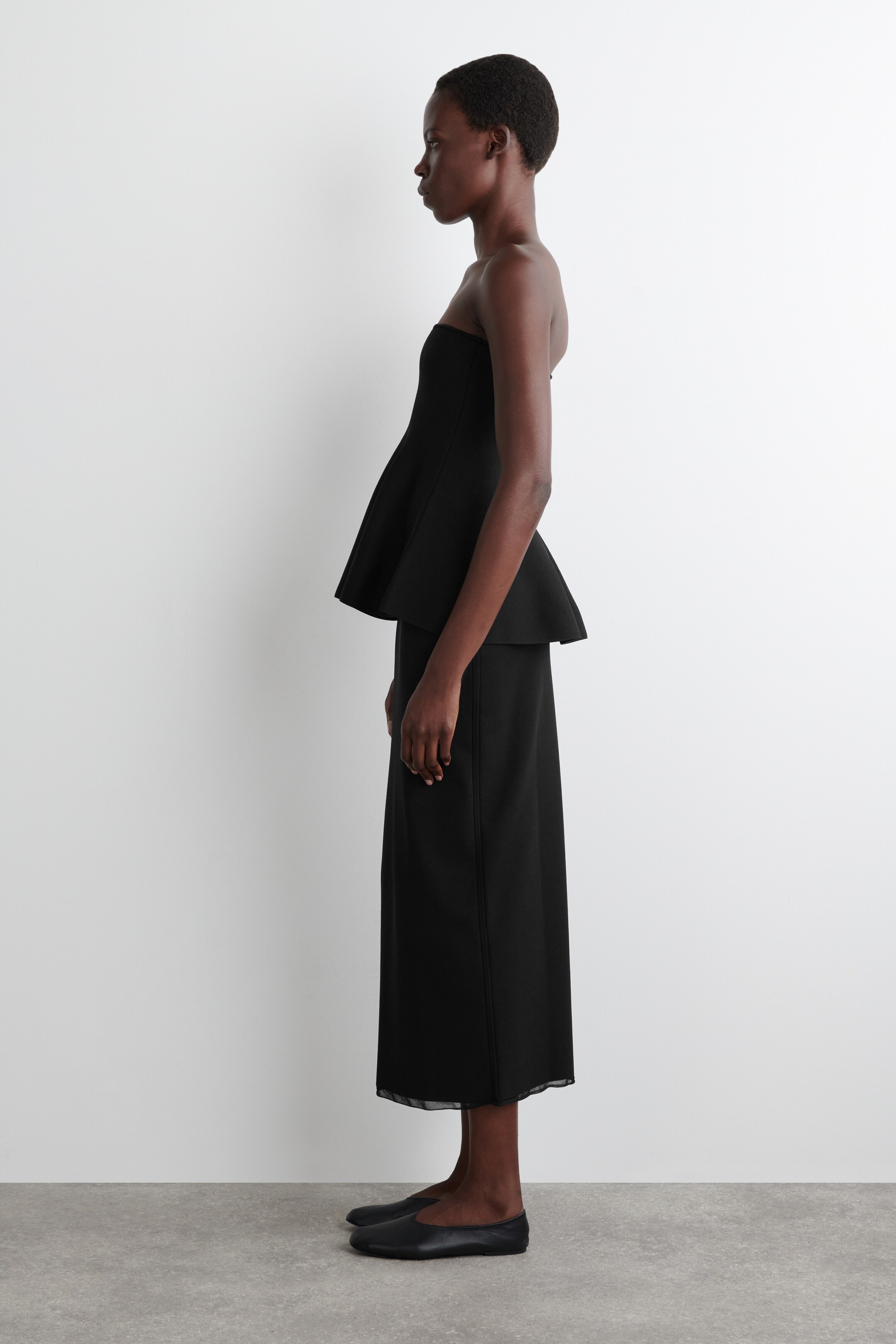 Ver imagen más grande: Un top negro sin tirantes con peplum se ensancha desde la cintura, combinado con una falda midi negra, visto desde un ángulo lateral. La falda cae en una silueta recta y presenta un dobladillo translúcido, con zapatos planos negros que completan el conjunto.