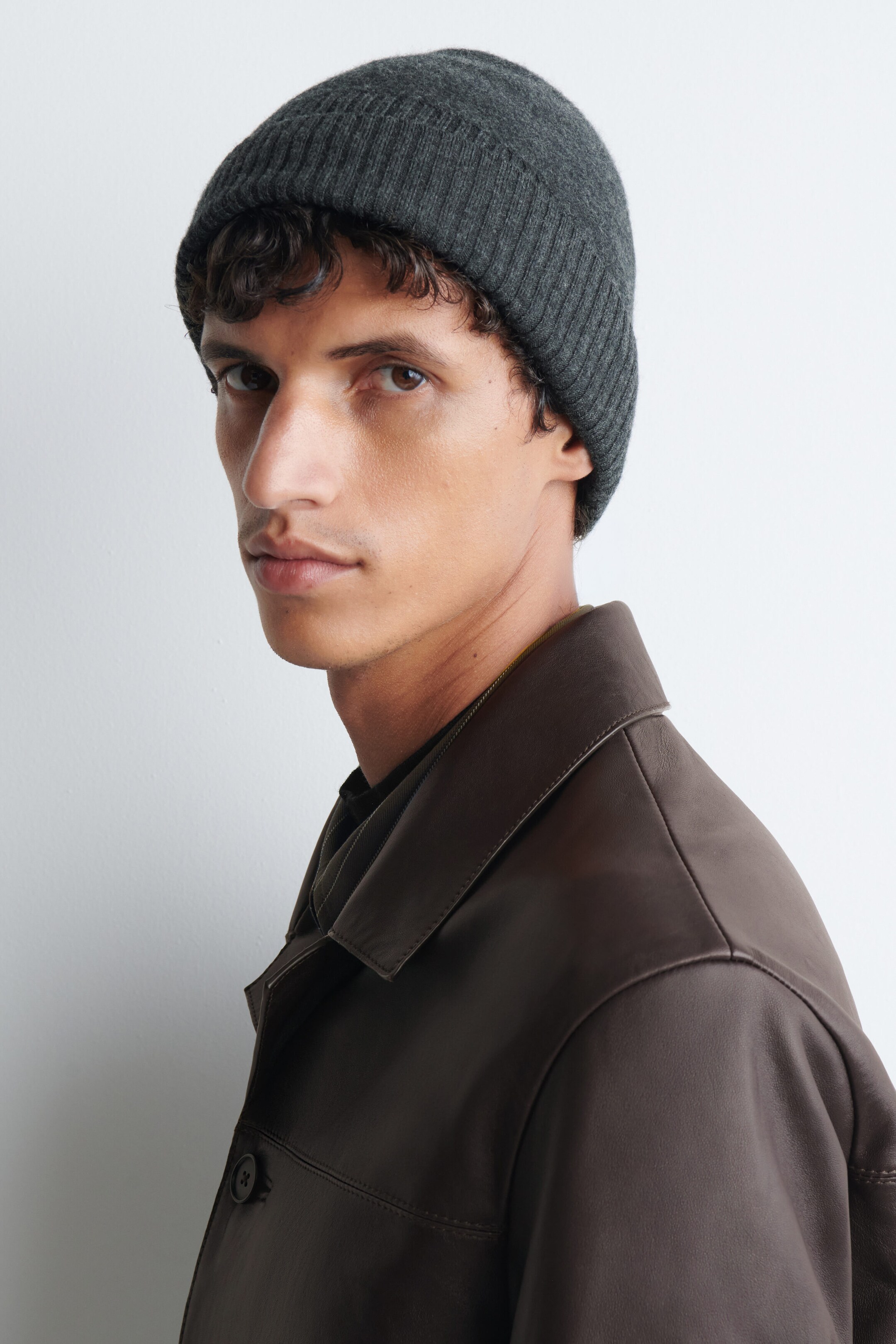 A dark grey knitted toque featuring a ribbed fold-up brim is being worn by the model. The toque is styled with a dark brown leather collared jacket.