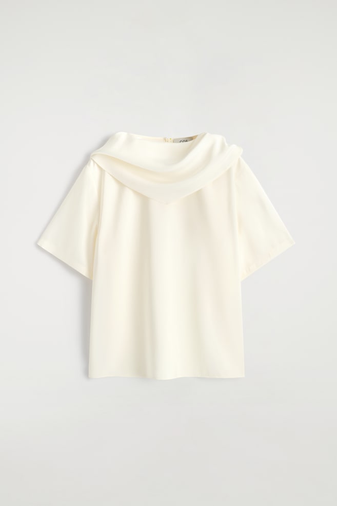 Una blusa color crema è mostrata dal retro, caratterizzata da maniche corte, un ampio scollo a cascata che drappeggia sulle spalle e una piccola cerniera di chiusura sul retro del collo. - 1