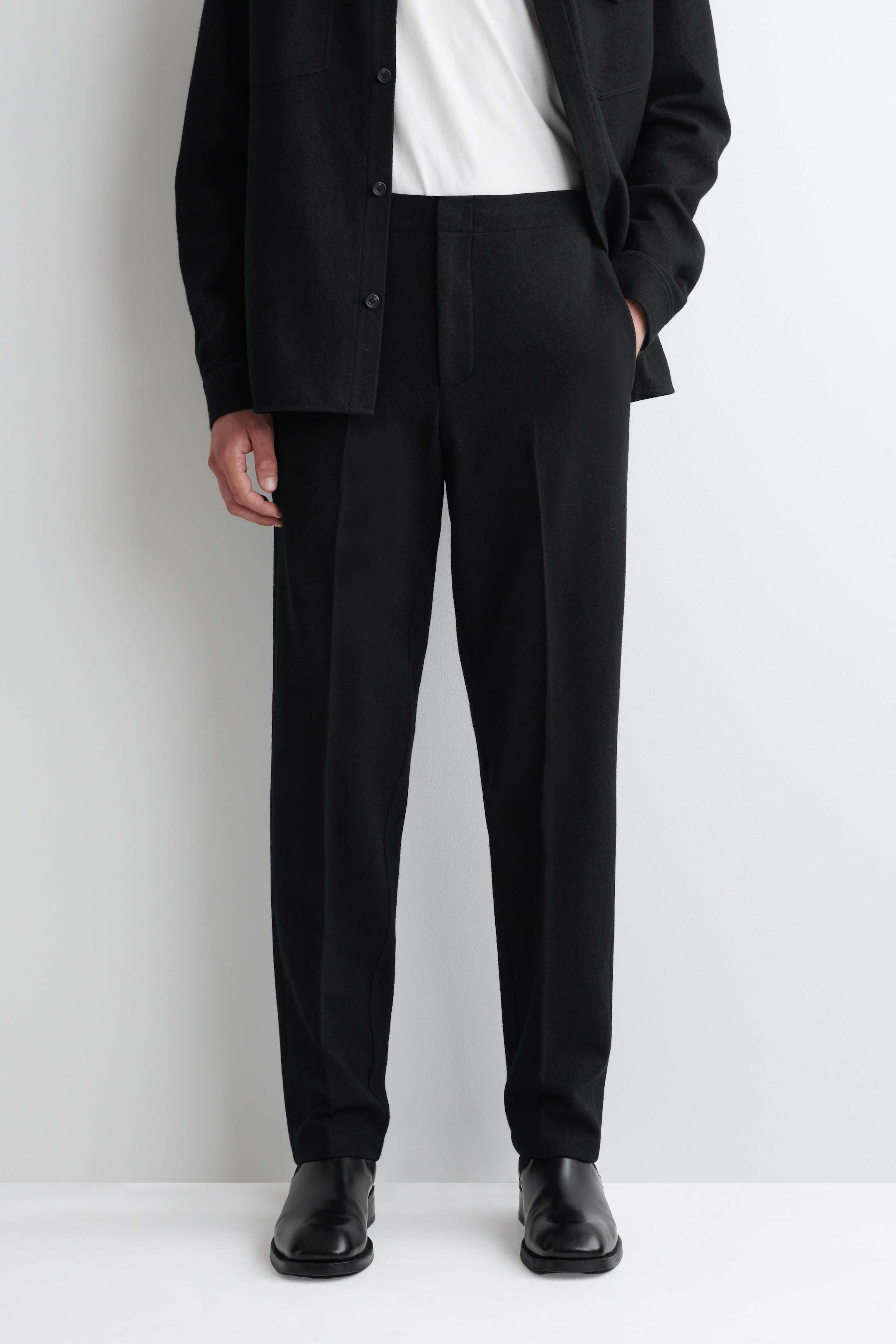 Agrandir l'image: Un pantalon noir est présenté de face, avec une coupe droite et décontractée et un design taille haute. Ils présentent un devant plat, une texture tissée subtile et des plis doux descendant le long de chaque jambe, avec des poches latérales visibles. Le pantalon repose sur des chaussures noires.