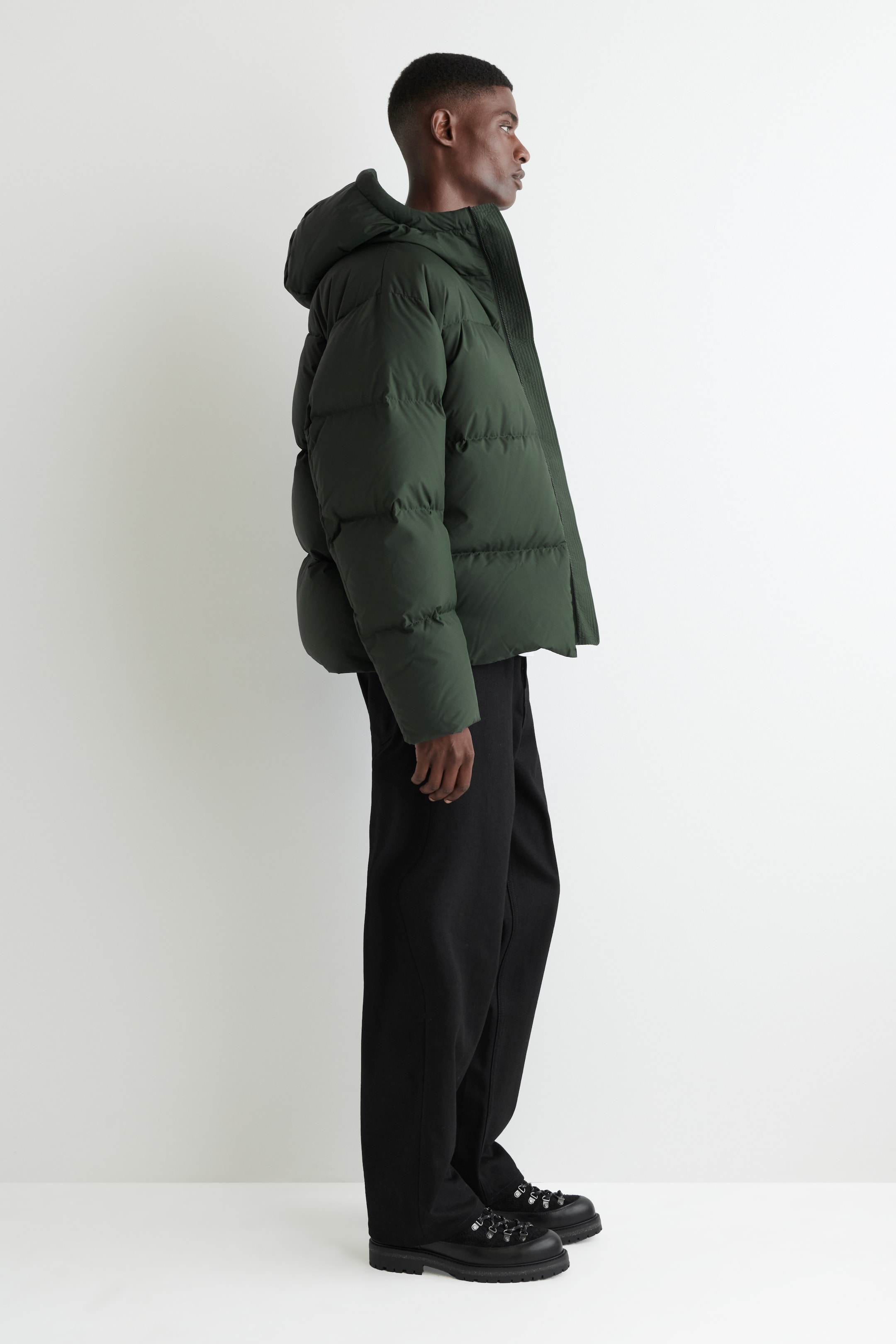 Ver imagem maior: Um casaco acolchoado verde escuro, de comprimento até à anca, está a ser usado de perfil lateral, exibindo o seu design volumoso com costuras horizontais distintas e um capuz anexado.