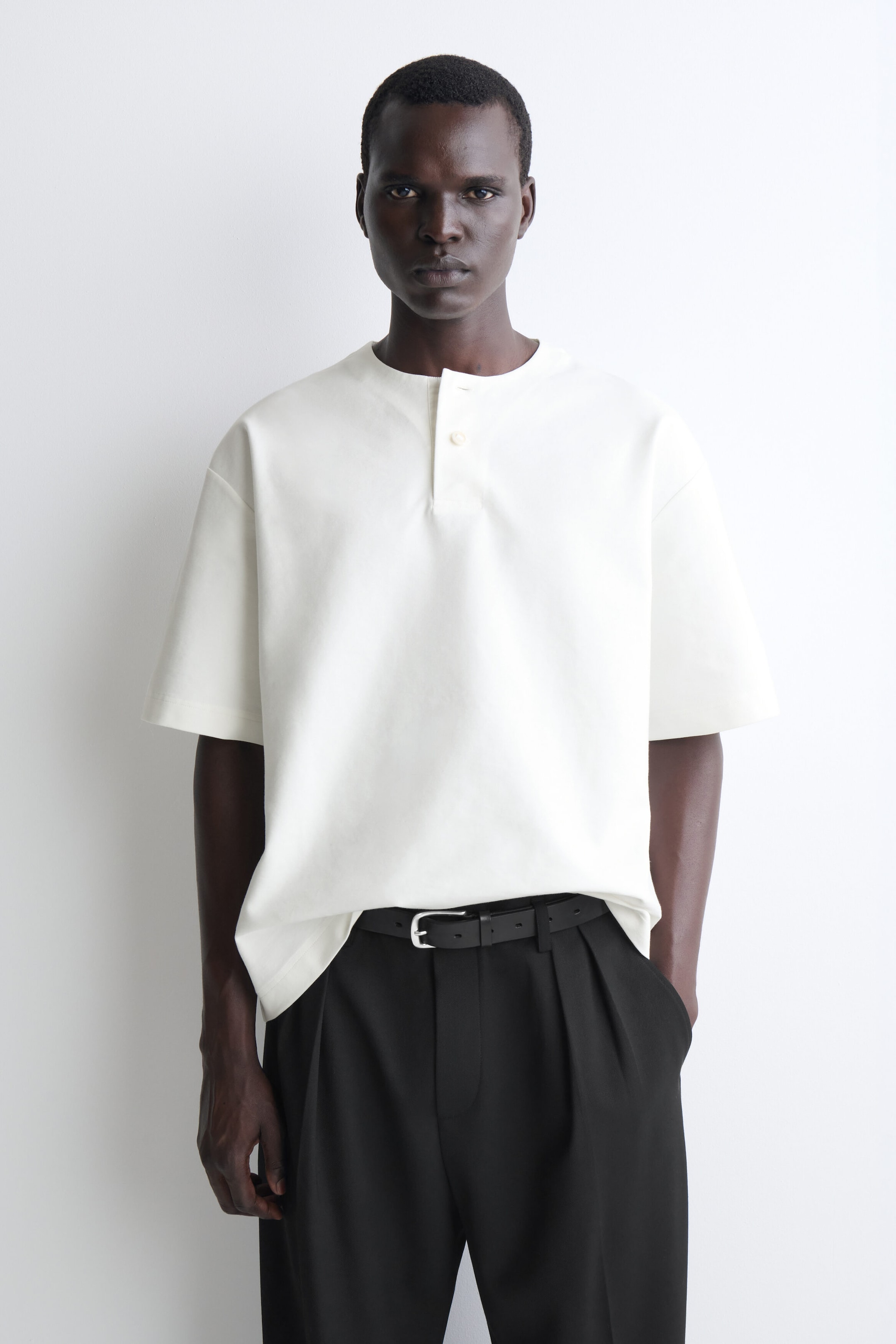 Ce t-shirt Henley surdimensionné en jersey de coton blanc cassé est présenté de face, avec un col rond, une patte de boutonnage partielle, des épaules tombantes et des manches larges à la longueur du coude. La coupe décontractée tombe amplement et le haut est partiellement rentré dans un pantalon noir avec une ceinture.