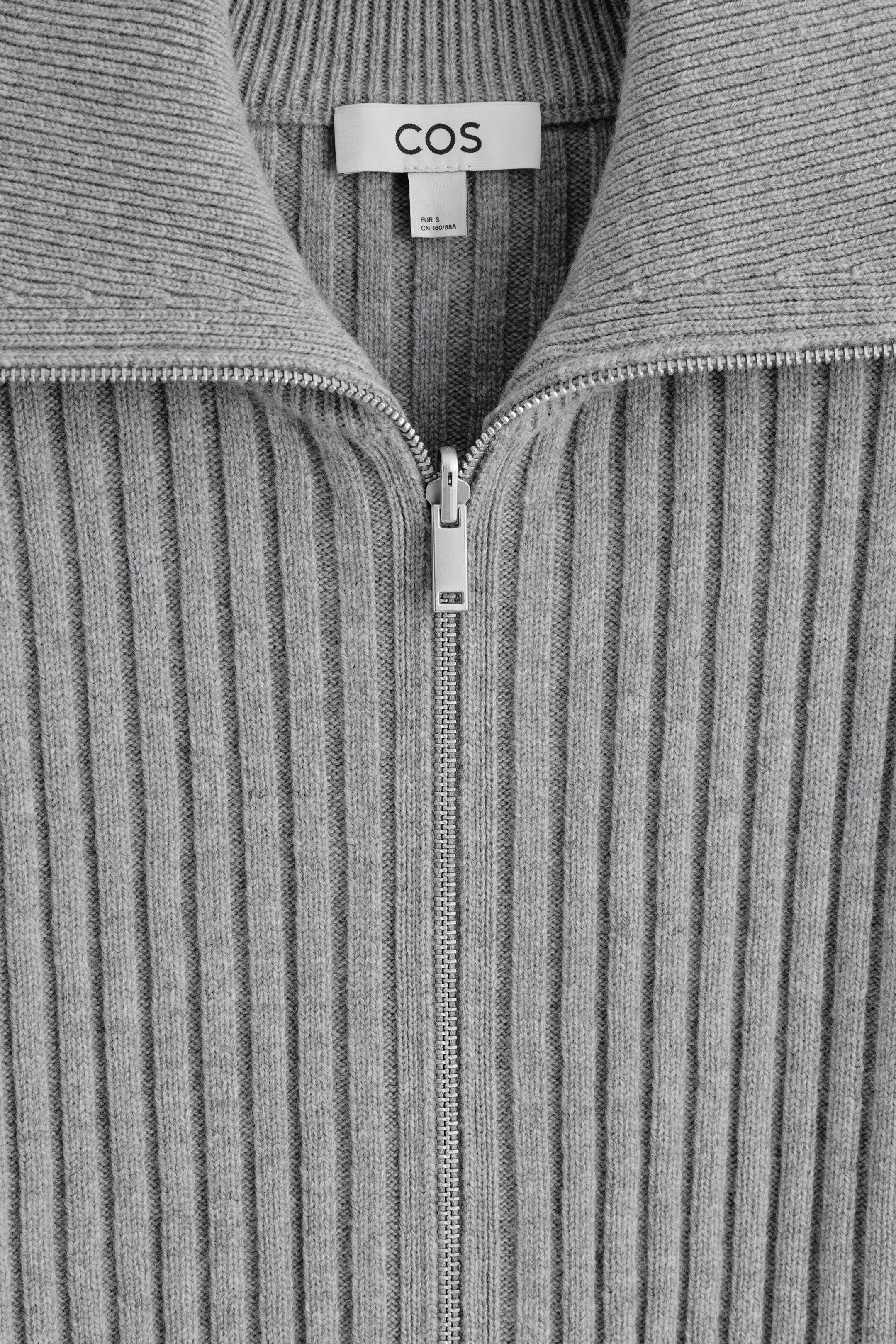 Größeres Bild ansehen: Ein grauer gerippter Pullover wird von vorne gezeigt, mit einem silberfarbenen Halb-Reißverschluss, der bis zu einem Stehkragen reicht. Ein weißes Markenetikett mit der Aufschrift „COS“ ist im Kragen sichtbar.