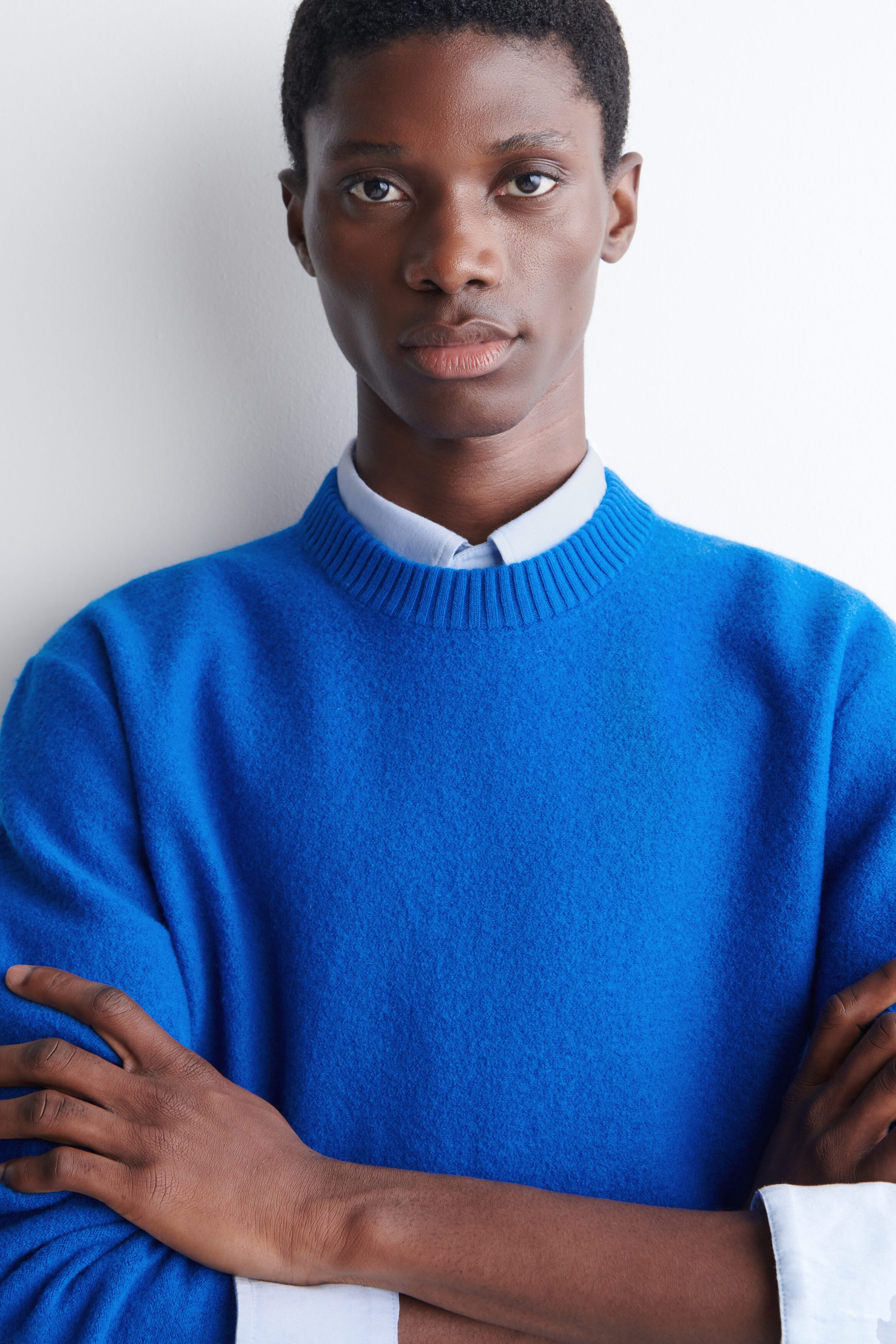 The model is wearing a bright blue crew-neck sweater layered over a light blue collared shirt. The sweater features a textured finish and a ribbed neckline. The model is wearing a bright blue crew-neck sweater layered over a light blue collared shirt. The sweater features a textured finish and a ribbed neckline.